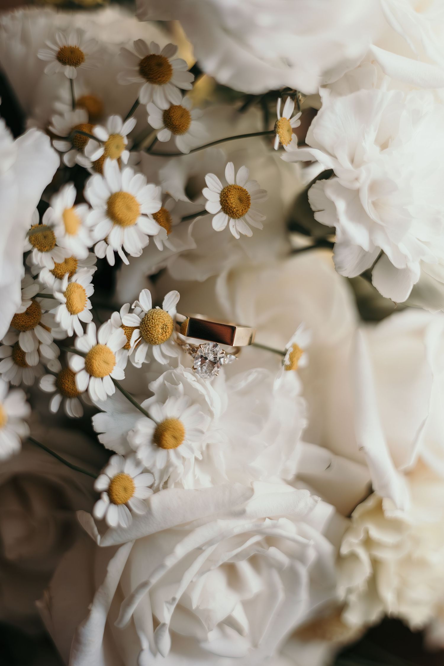 Gold diamond engagement ring resting on white roses and daisies in a bridal bouquet.