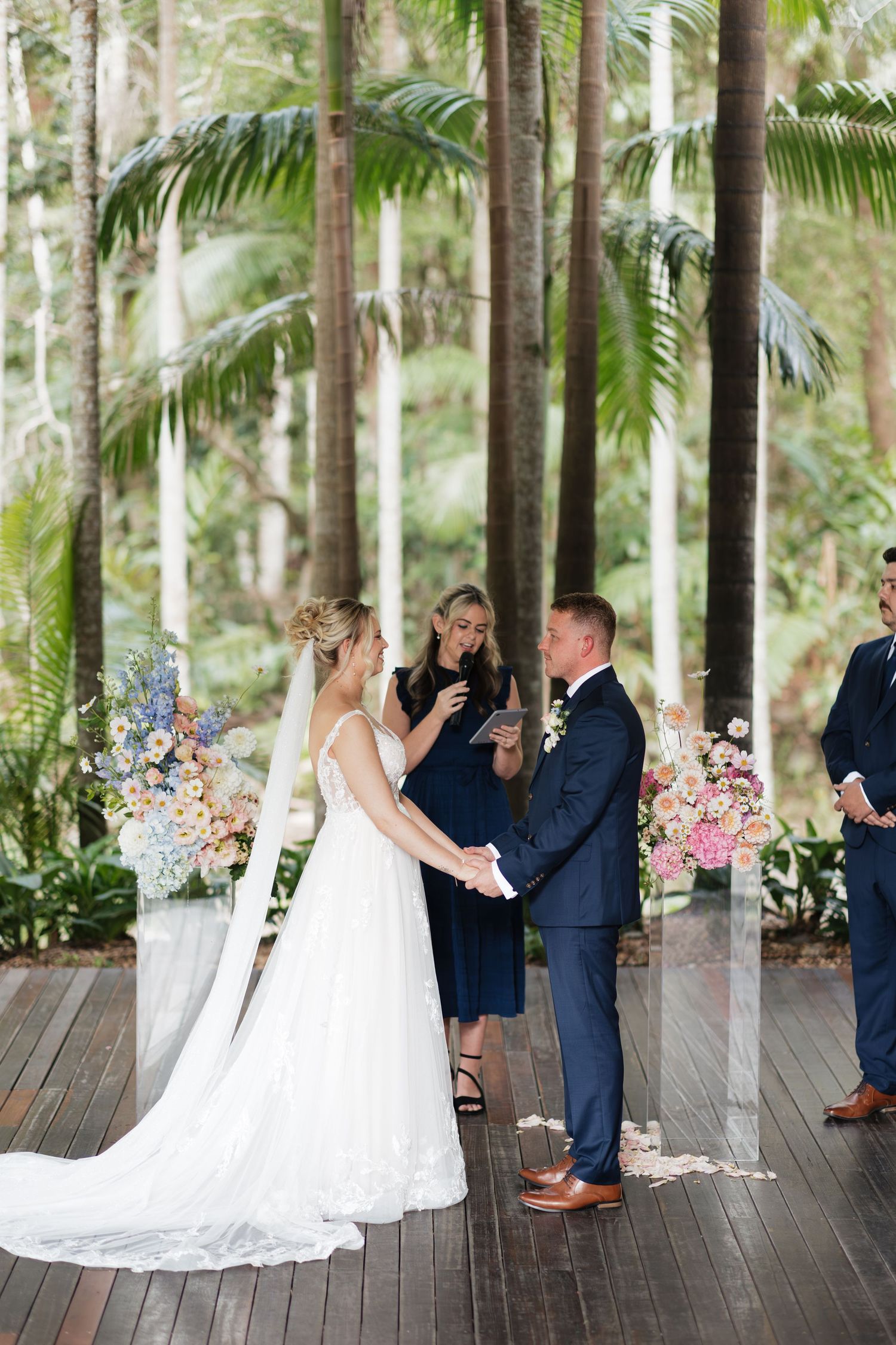 Bride and groom exchange vows at an outdoor wedding ceremony surrounded by tropical palms and colorful floral arrangements.