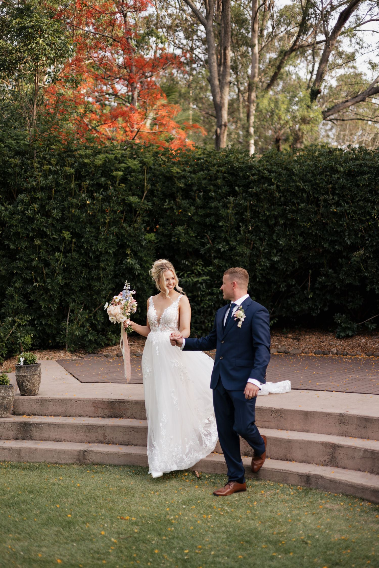 Bride and groom walking hand in hand through lush garden grounds, bride holding bouquet in elegant white gown.