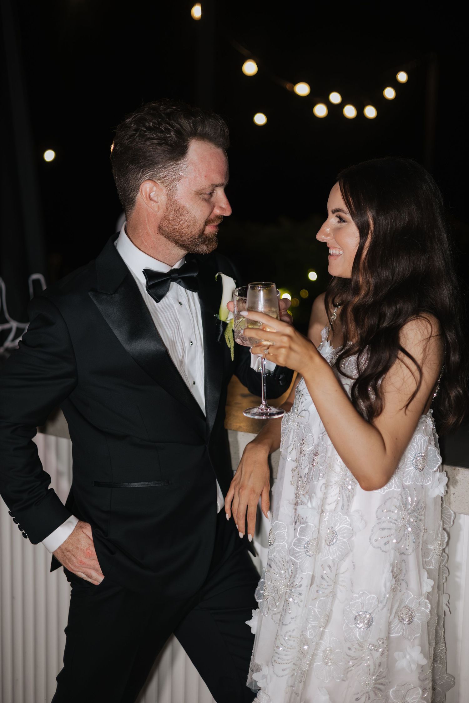 Bride and groom in formal attire toasting with champagne glasses at an elegant outdoor evening wedding reception.