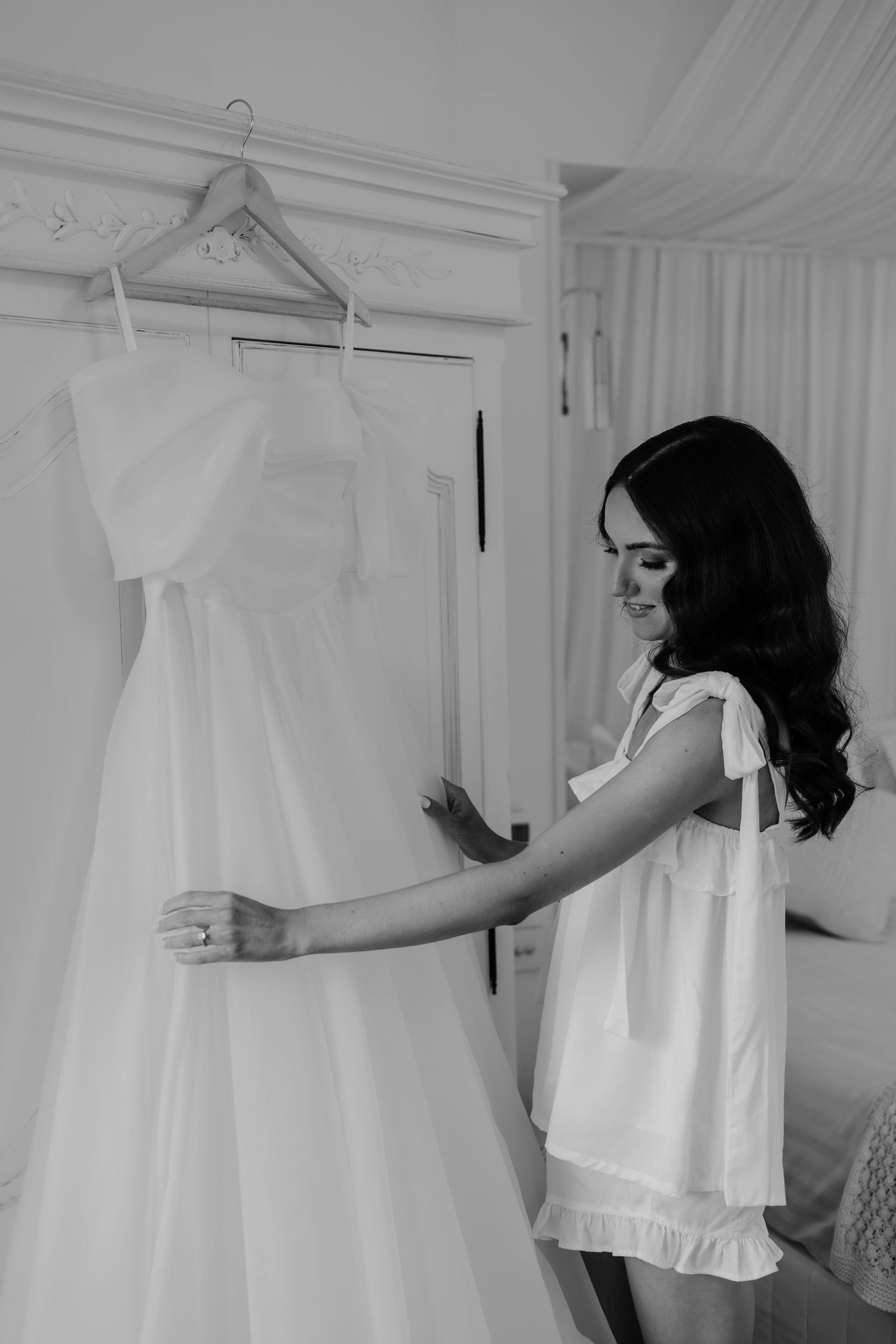 Bride in white robe admiring her wedding dress hanging on door in black and white photo.