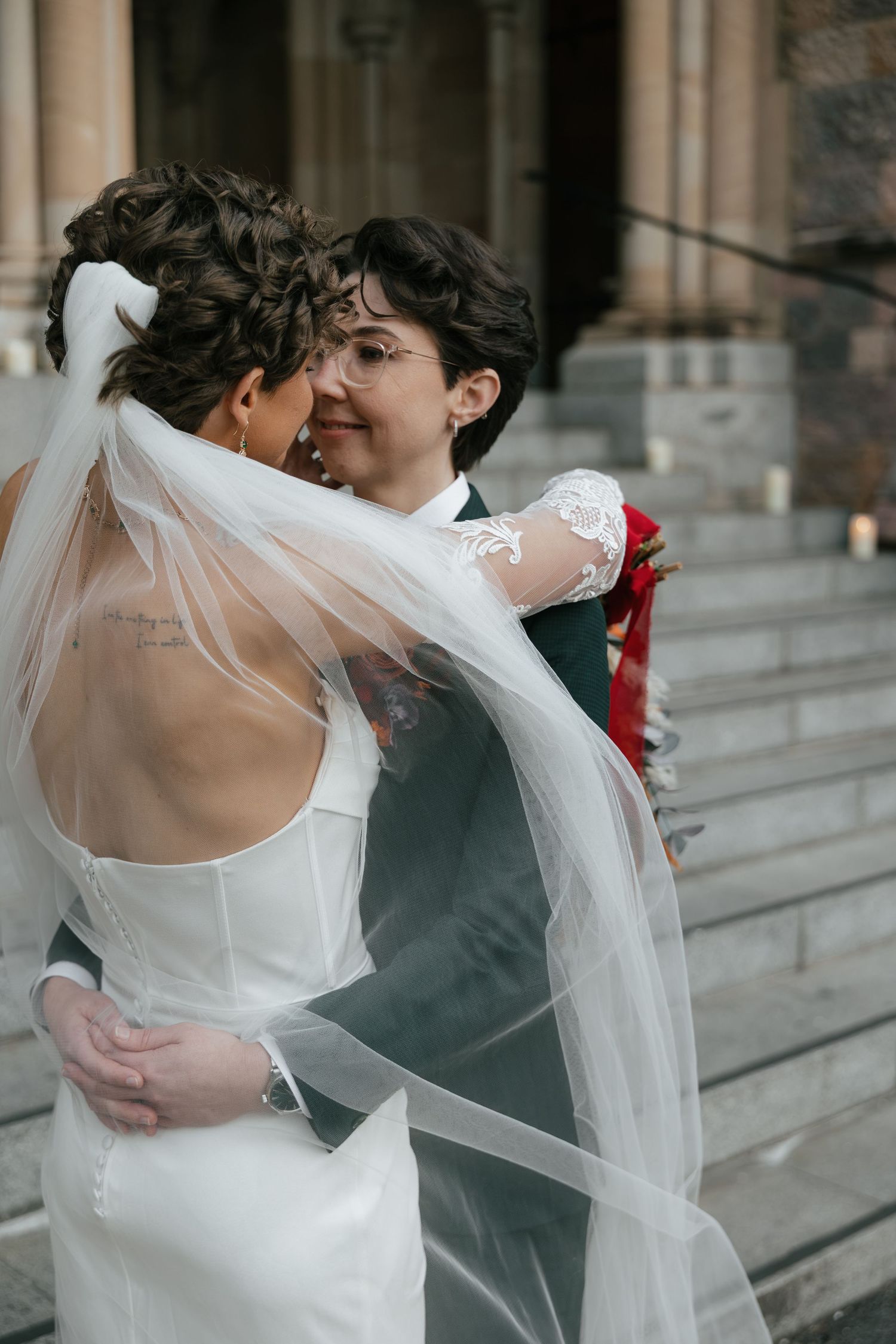 Two brides embrace on church steps, one in a white gown with veil, the other in a suit with lace sleeves.