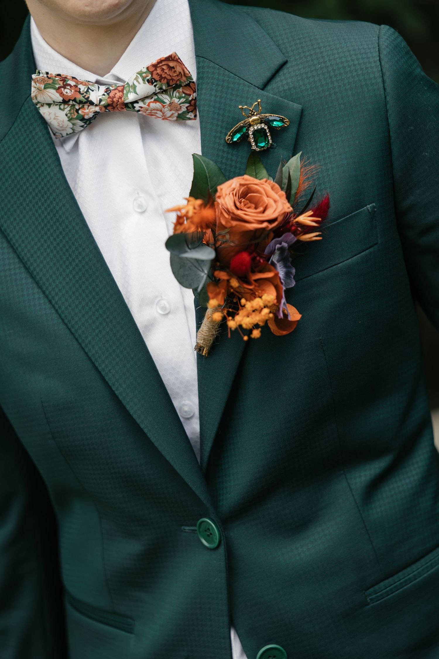 Groom in green suit with floral boutonniere featuring orange rose, colorful bow tie, and decorative bee brooch.