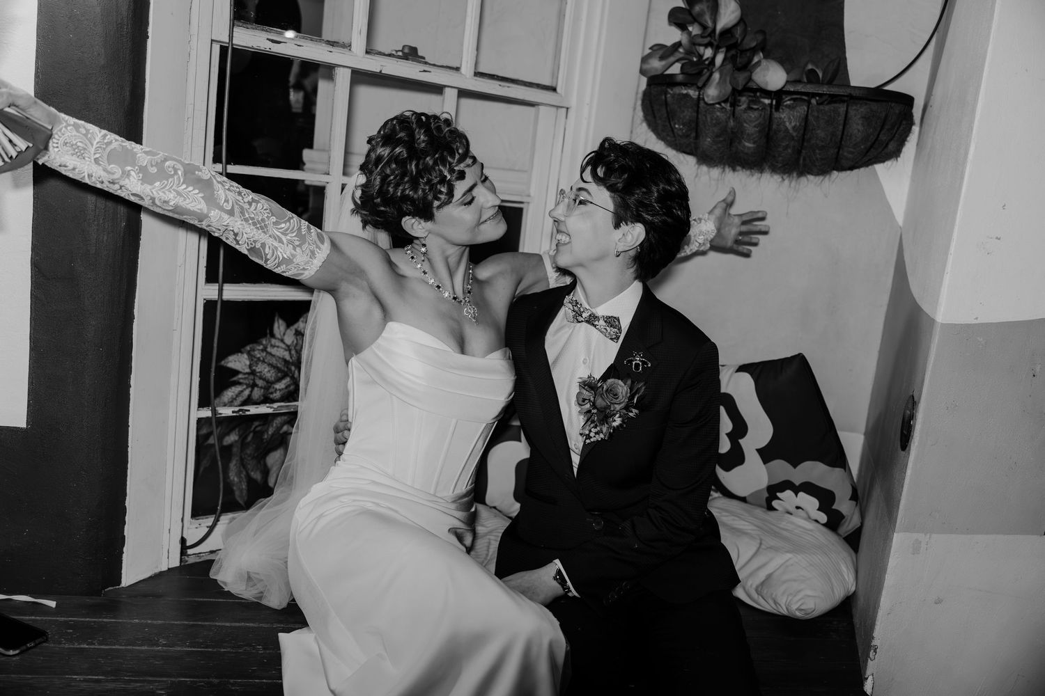 Black and white photo of two brides sharing a joyful moment, one in a strapless gown with tattoos, at their wedding.