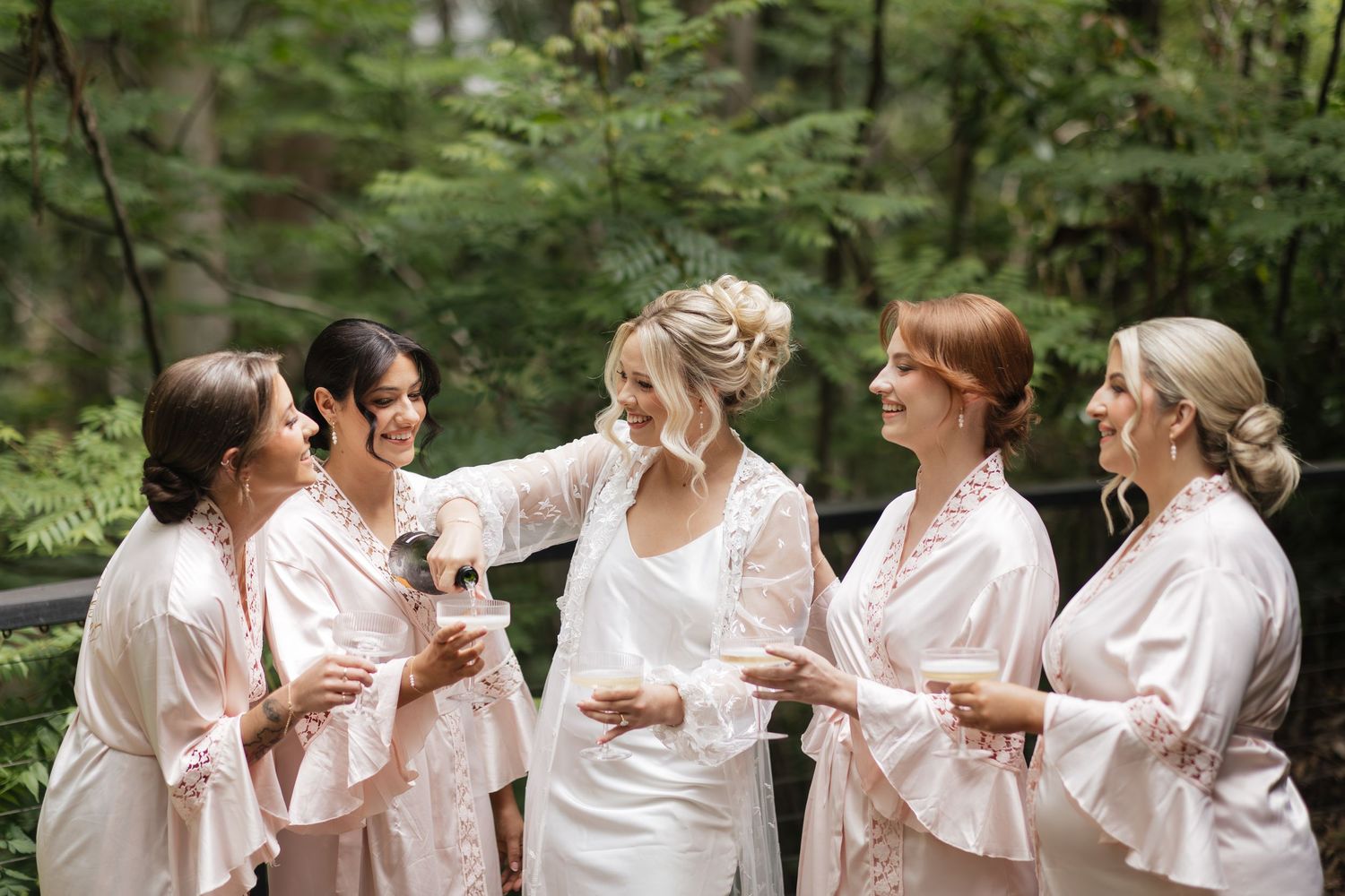 Bride in white robe laughing with four bridesmaids in pink robes holding coffee cups on a lush green outdoor deck.