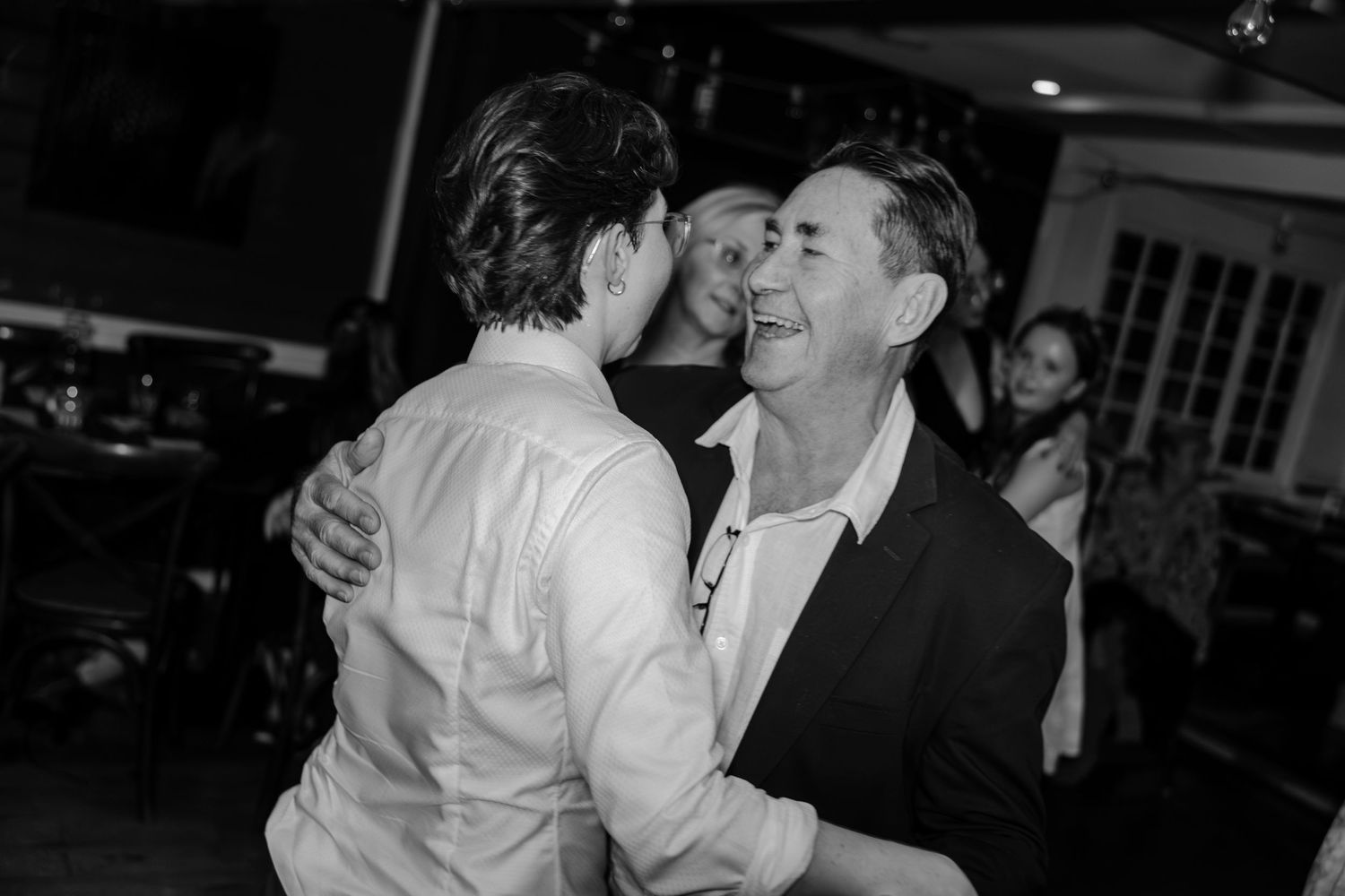 Black and white photo of two men embracing and laughing joyfully on a dance floor at a social event.