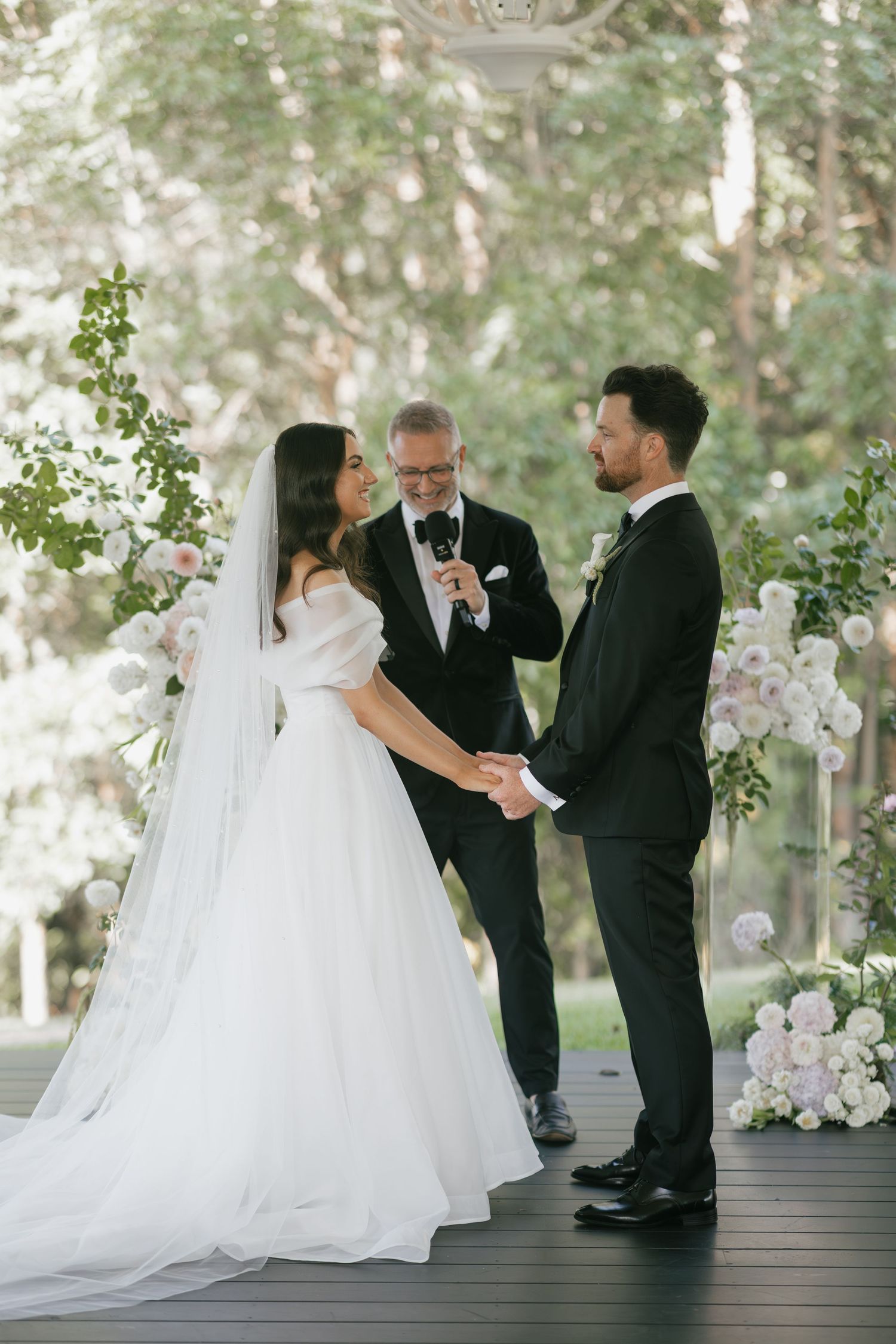 Bride and groom holding hands during outdoor wedding ceremony with floral arch and officiant in forest setting.