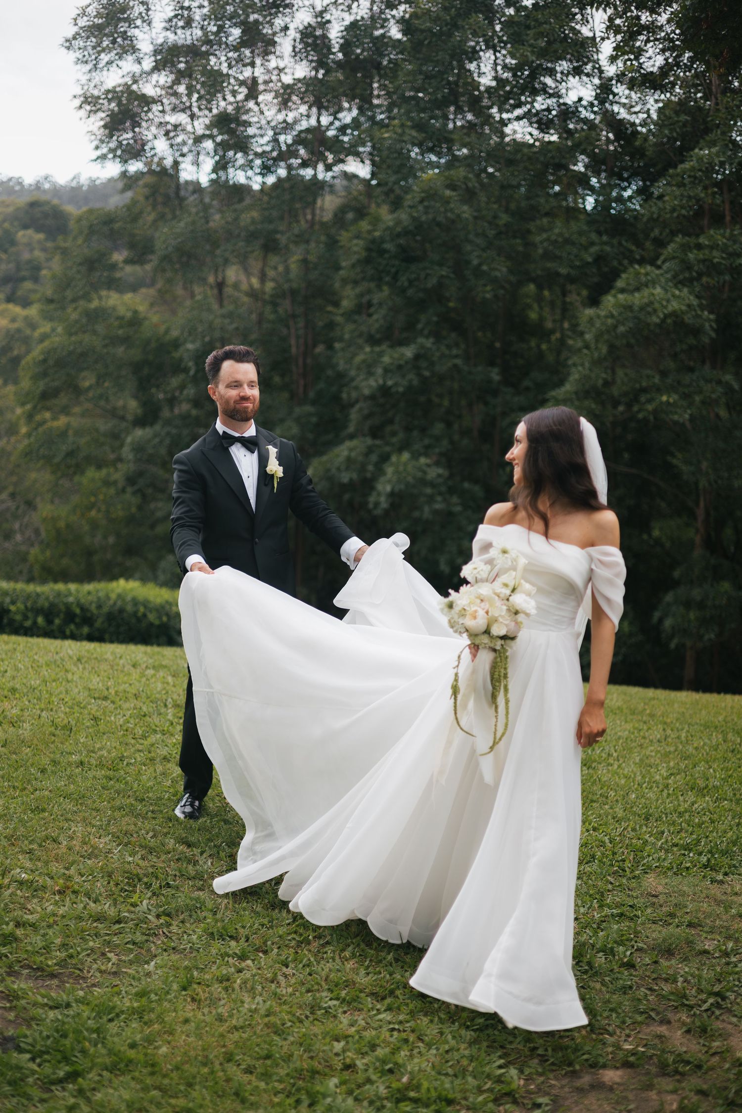 Bride in white off-shoulder gown and groom in black tuxedo dancing on green lawn with lush forest backdrop.