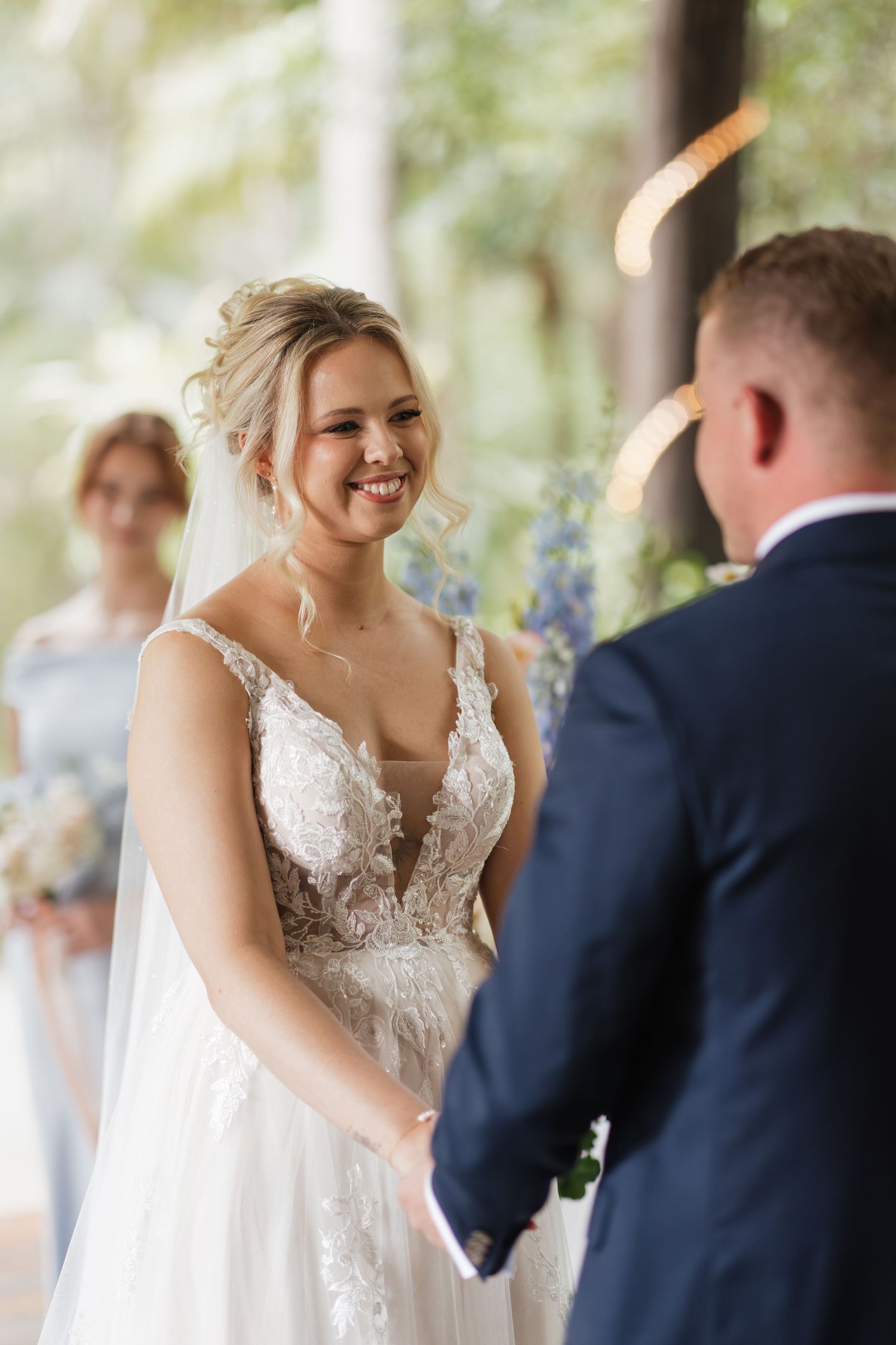 Smiling bride in lace wedding gown holds hands with groom during outdoor wedding ceremony with bridesmaid in background.