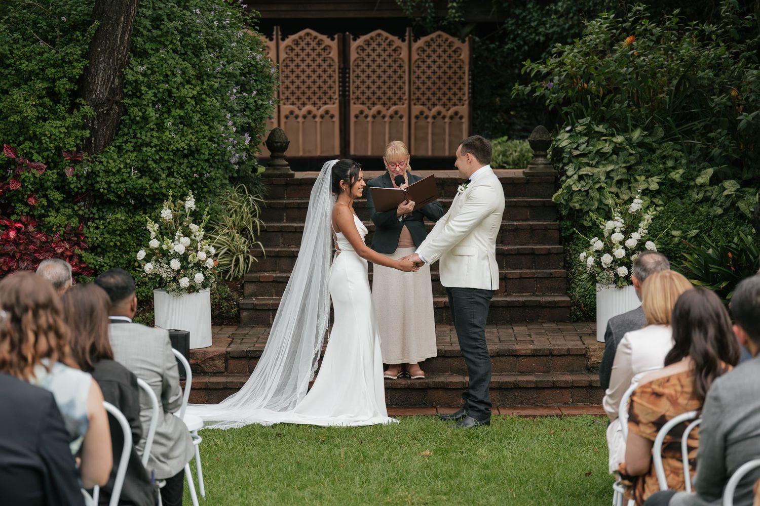 Outdoor wedding ceremony with bride and groom holding hands before officiant, flanked by white floral arrangements on lush garden steps.