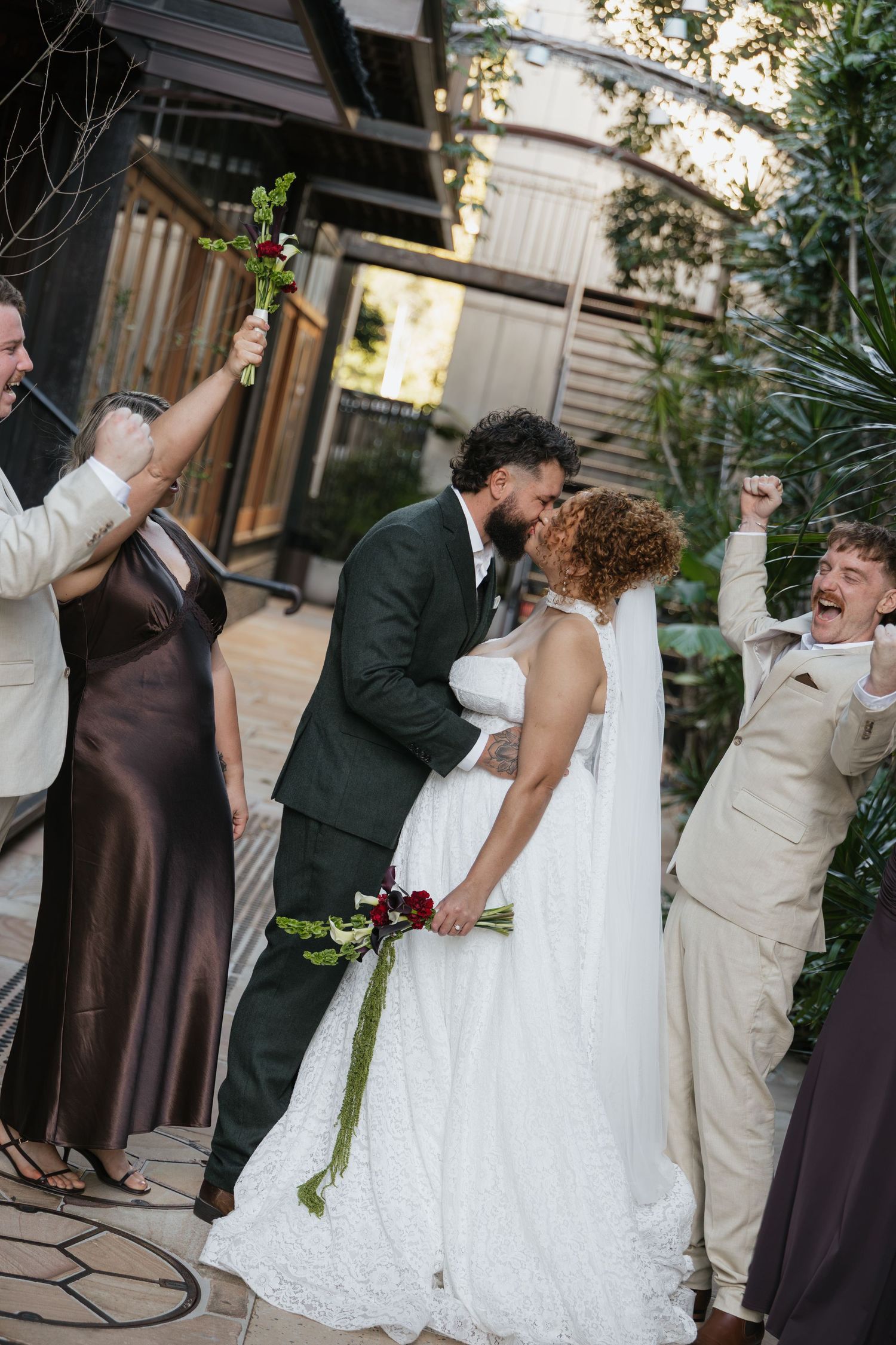A joyful wedding couple shares their first kiss outdoors, surrounded by cheering guests, bride holding a green bouquet.
