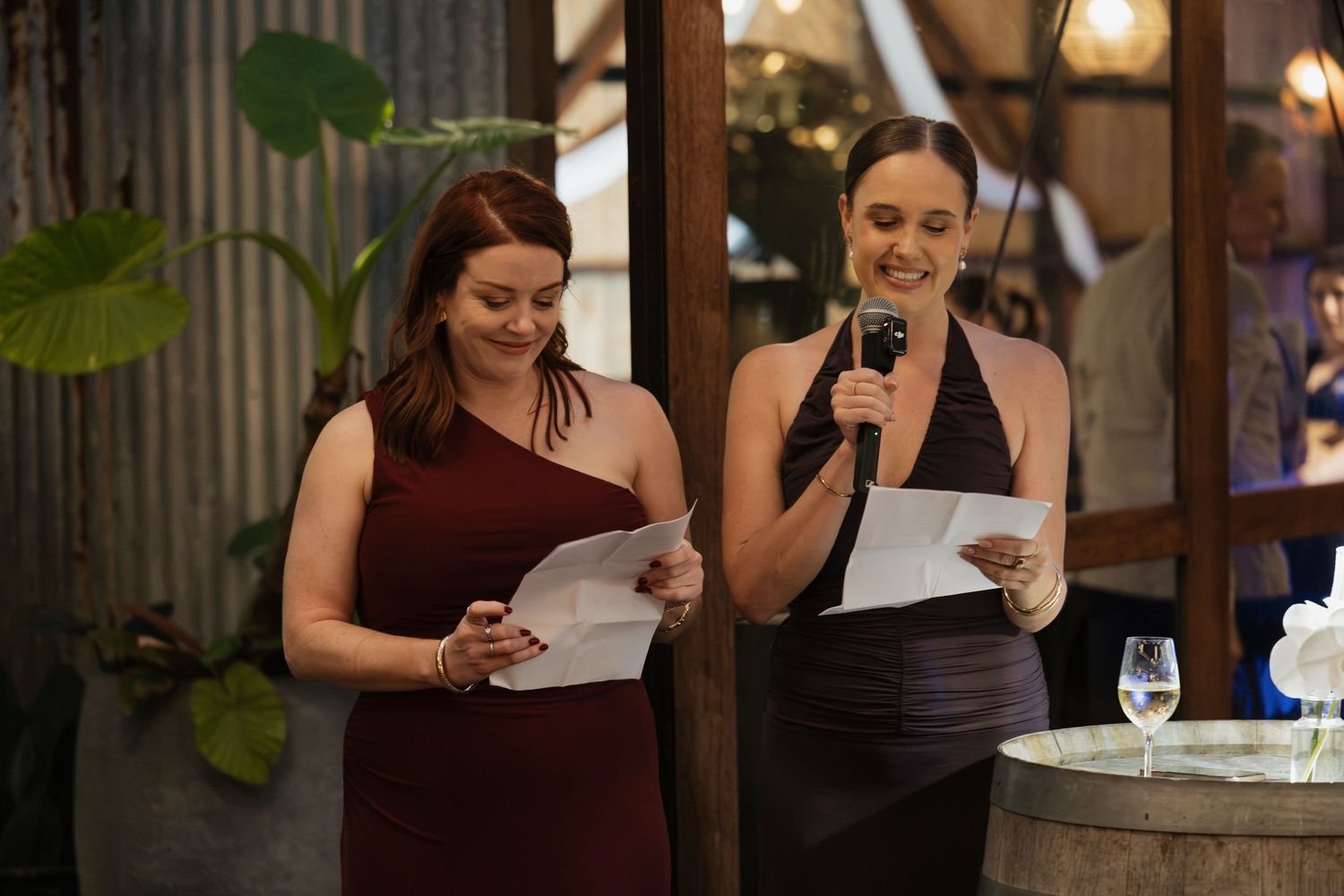 Two bridesmaids in elegant dresses delivering a speech at a wedding reception, holding papers and a microphone.