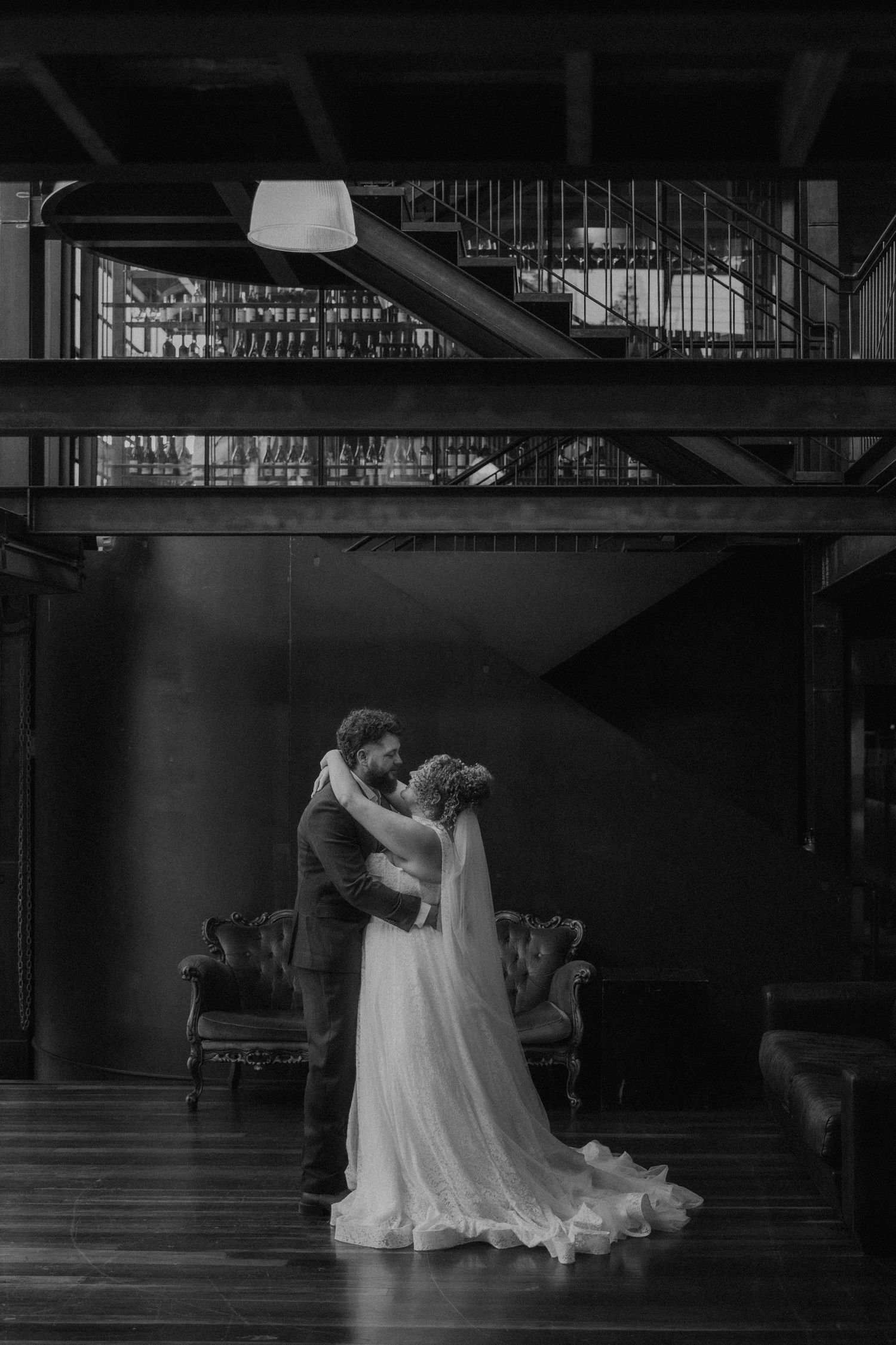 Black and white photo of a bride and groom embracing in an elegant industrial venue with a staircase backdrop.