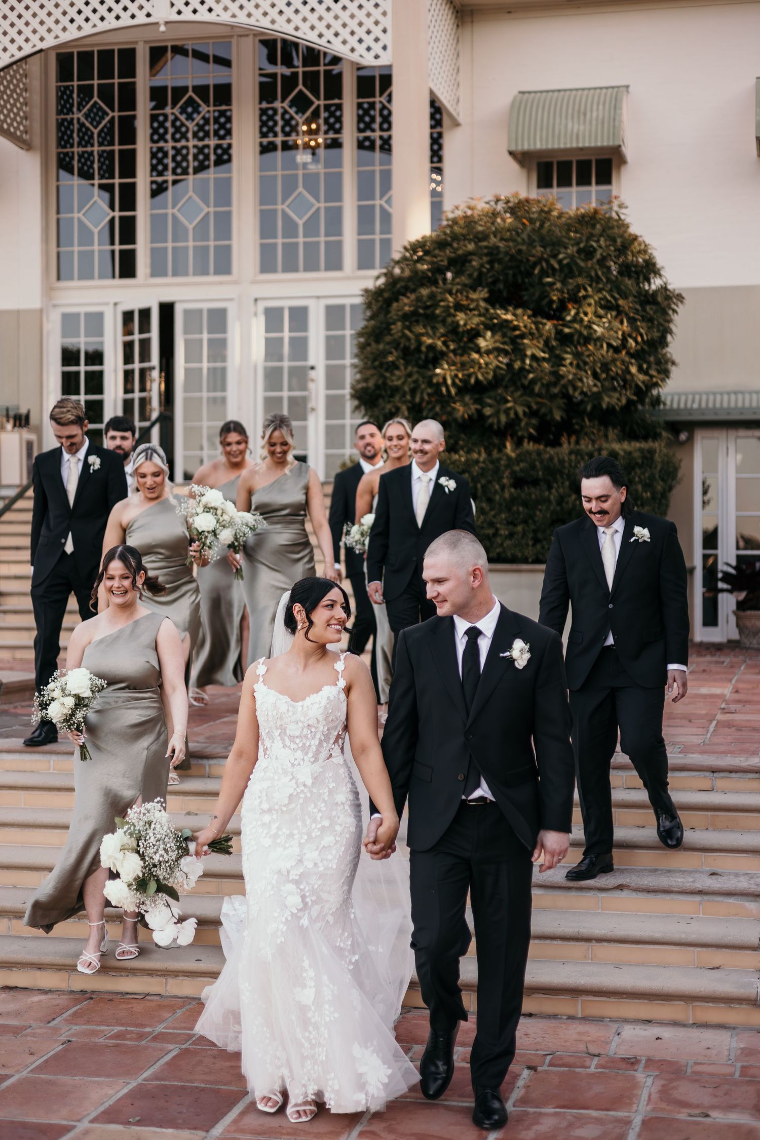 Wedding party members walk down outdoor steps at an elegant venue with decorative railings and archways.