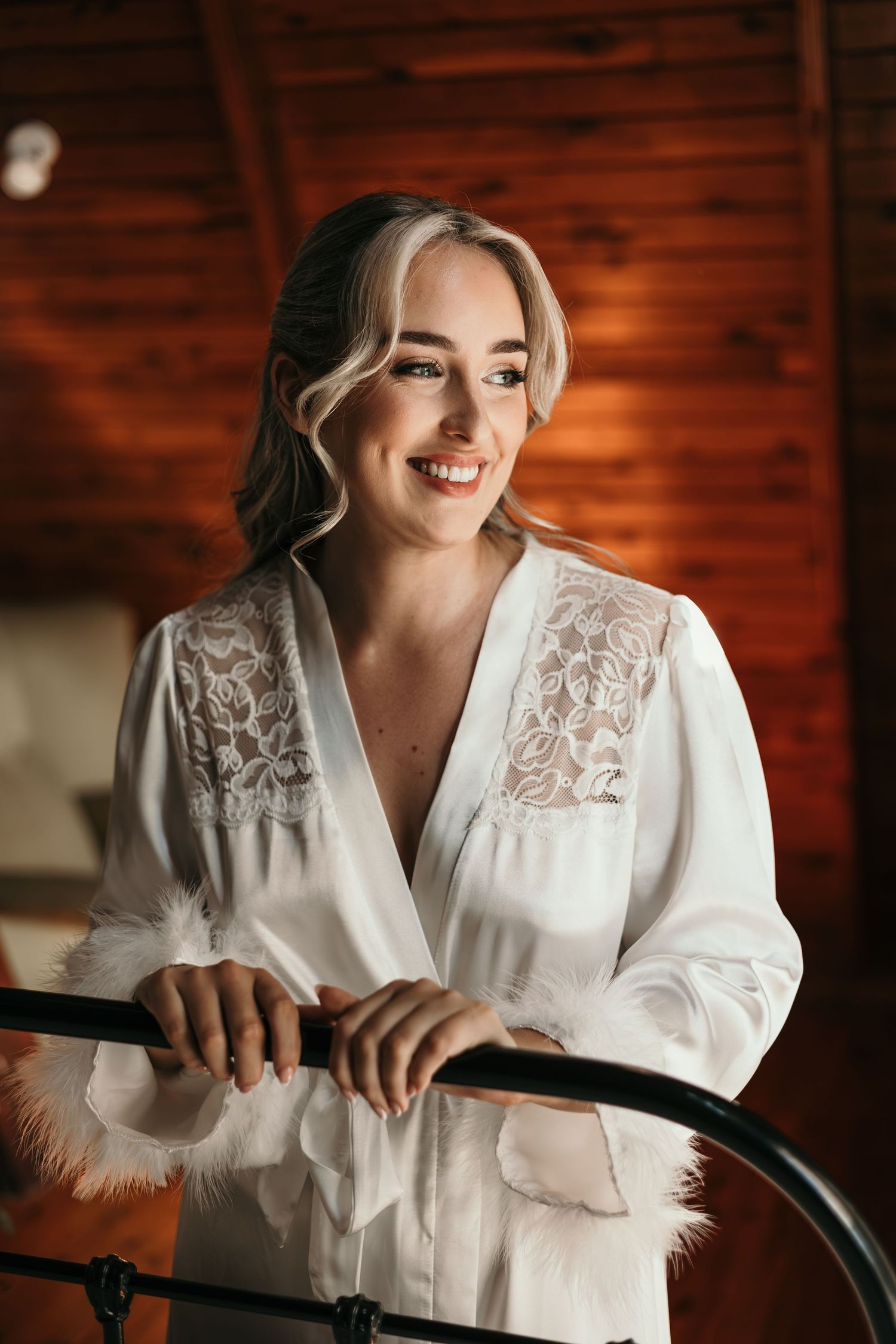 Smiling blonde bride in white lace satin robe leaning on black bed frame in warm wooden interior.