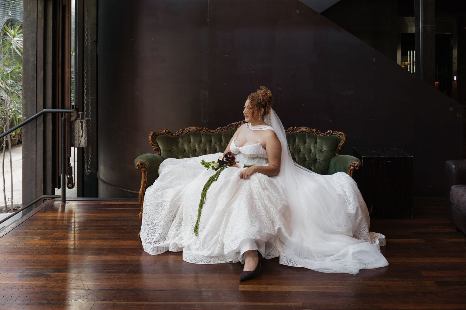 Bride in white wedding gown sitting on vintage green velvet sofa, holding bouquet, in elegant dark wood interior.
