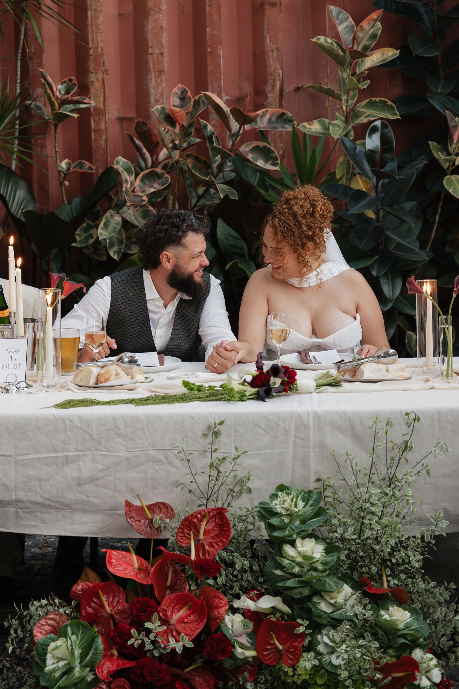 Bride and groom sitting together at elegant wedding reception table with red floral arrangements and tropical greenery backdrop.