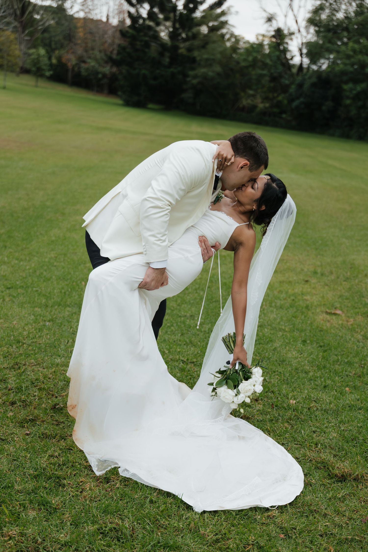 Groom in white suit dips bride in wedding dress for a romantic kiss on green lawn outdoors.