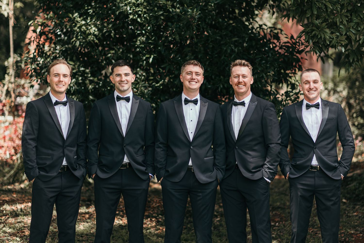 Five groomsmen in matching black tuxedos with bow ties posing outdoors in front of lush green trees.