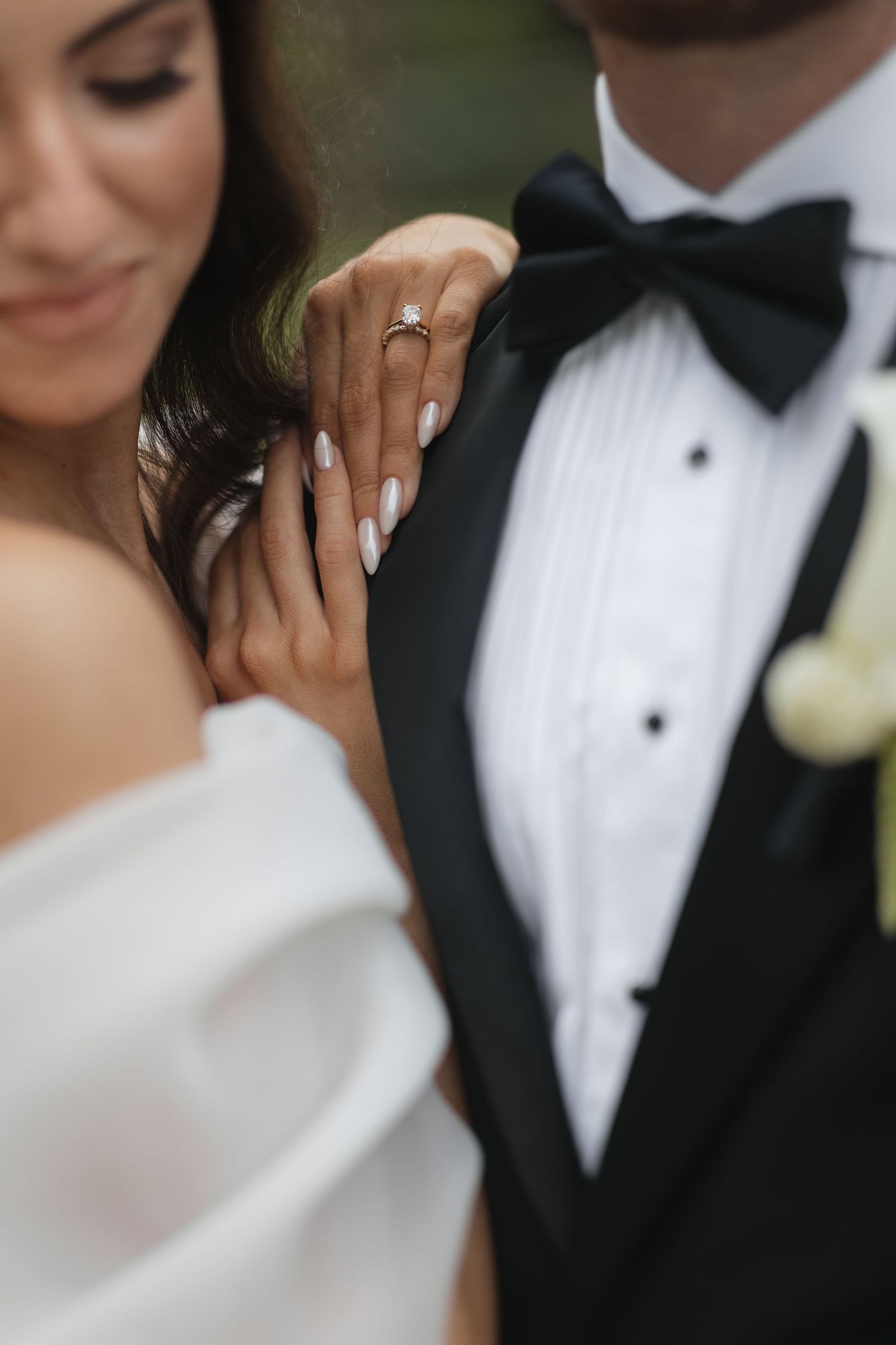 Bride in off-shoulder gown displays diamond engagement ring while holding groom's arm in black tuxedo.
