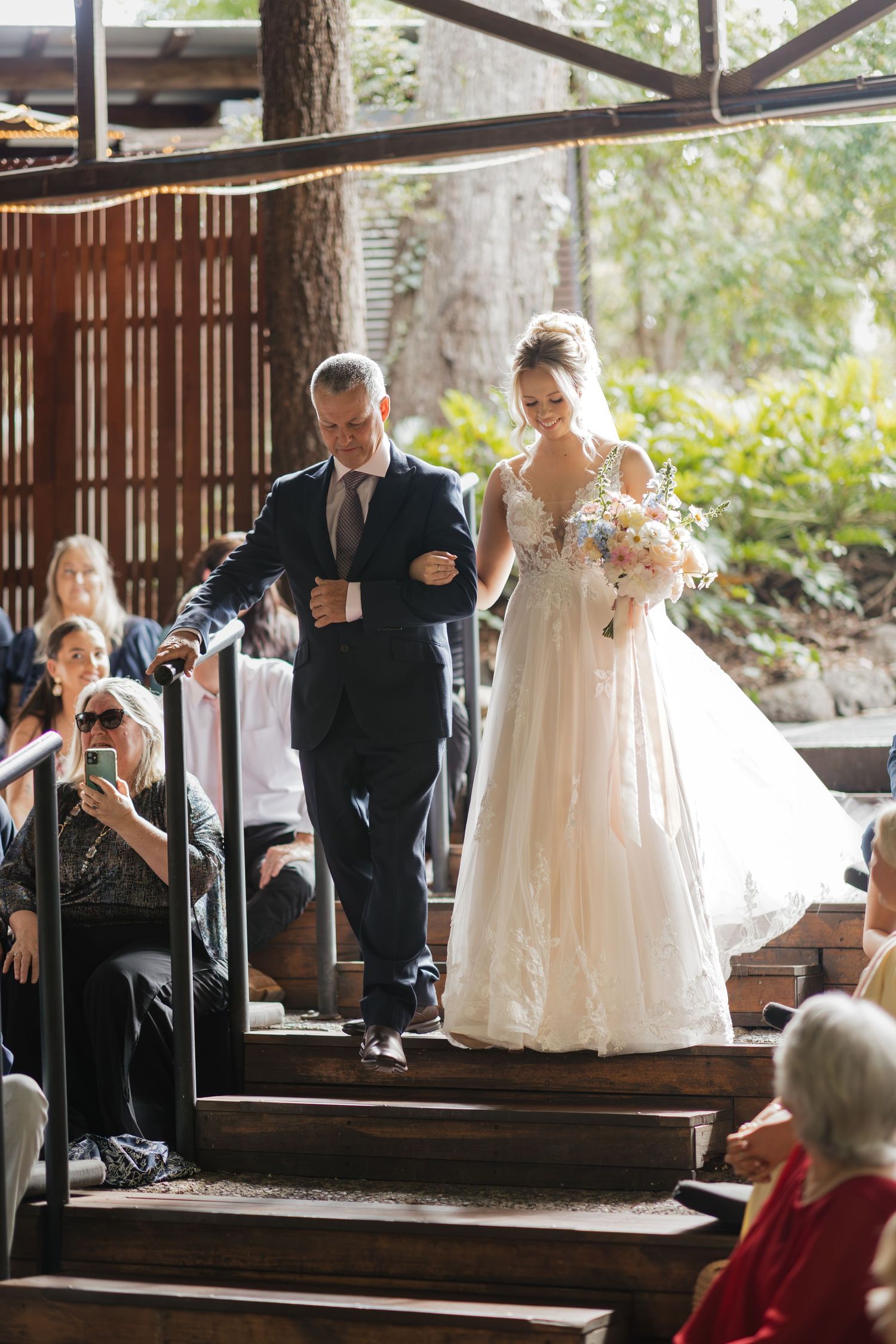 Bride in white gown walks down wooden steps with escort at outdoor wedding ceremony.
