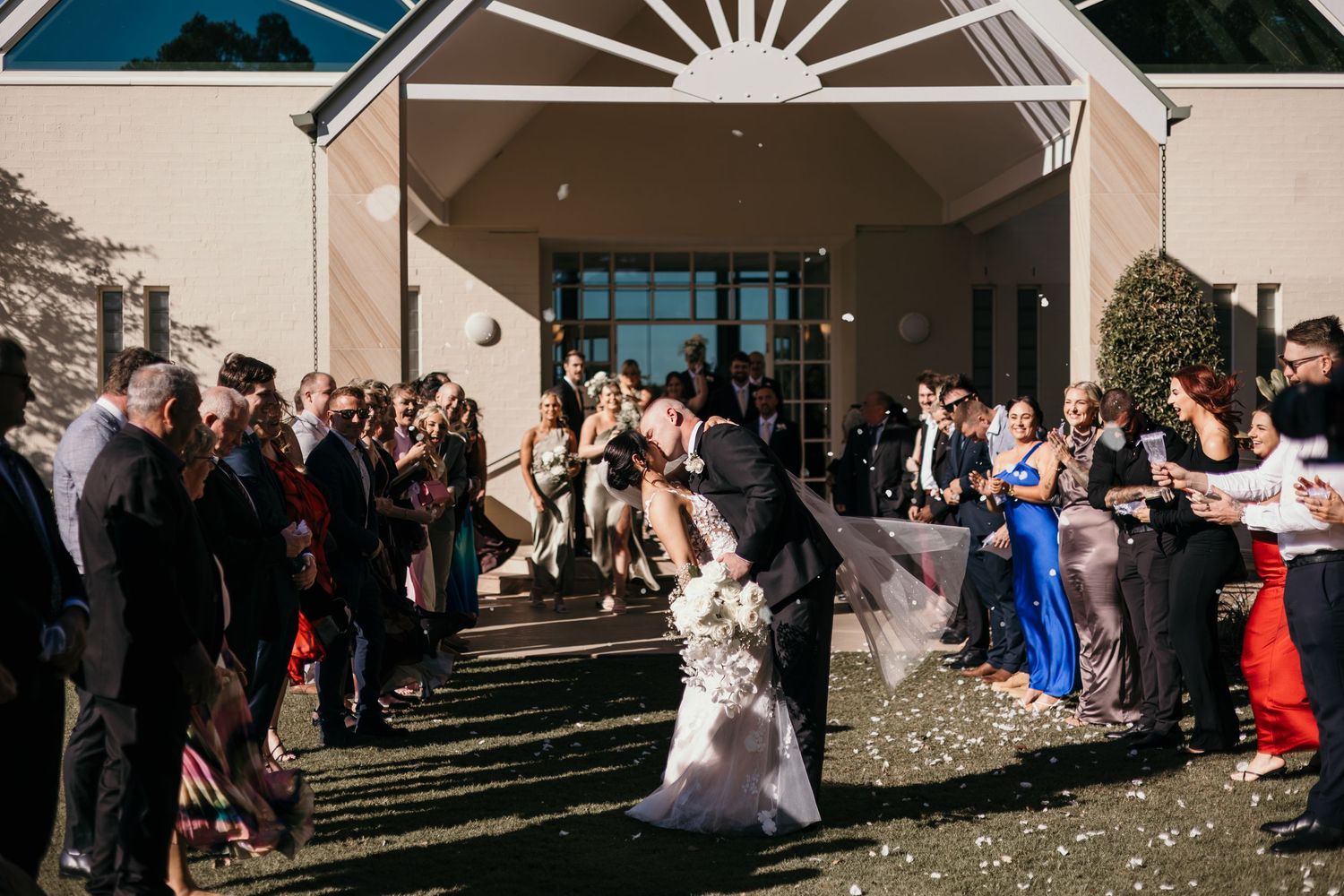 Wedding guests toss white flower petals as newlyweds exit their ceremony on a sunny afternoon.