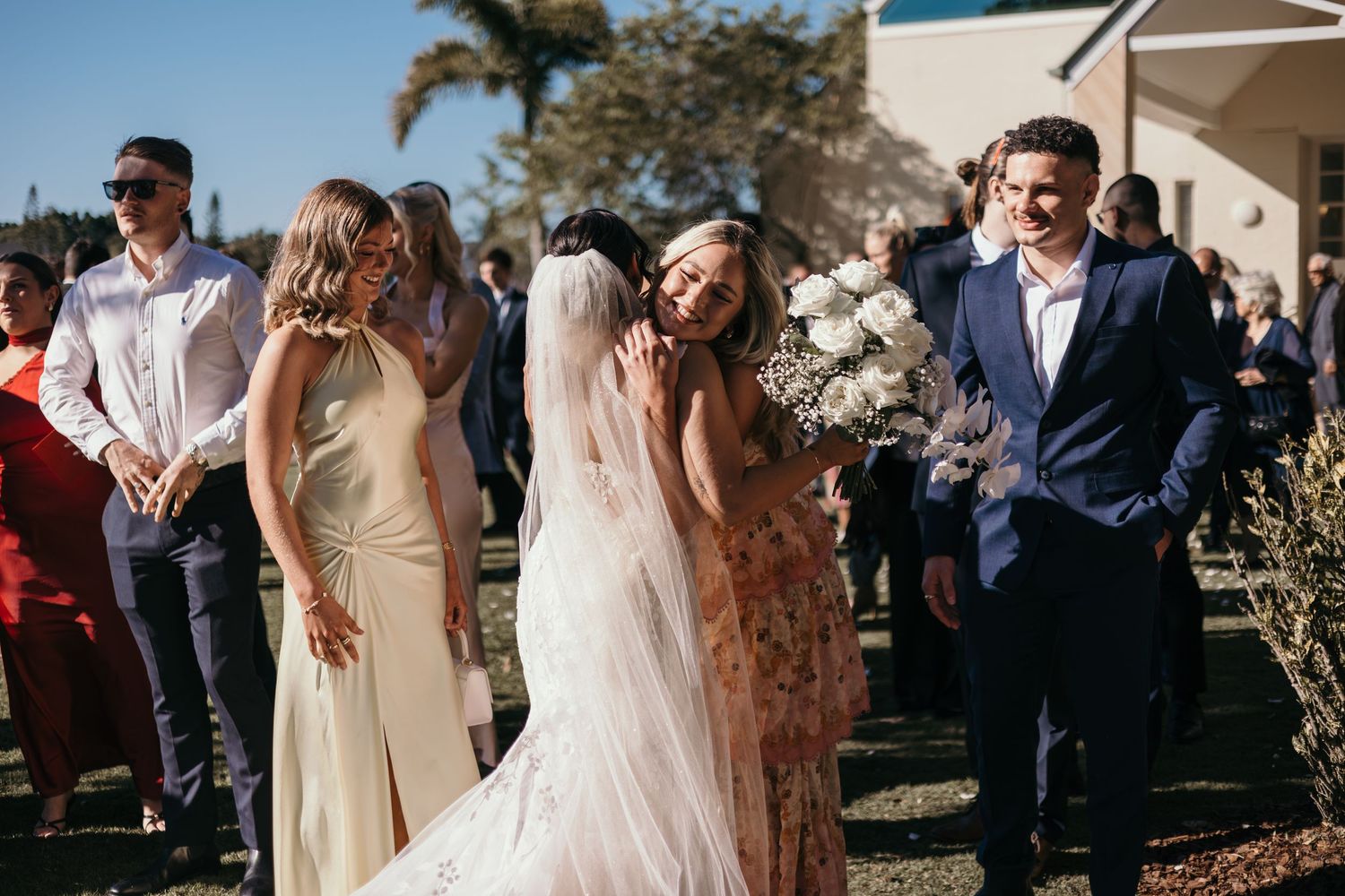 Guests celebrate at an outdoor wedding reception on a sunny day with palm trees in the background.