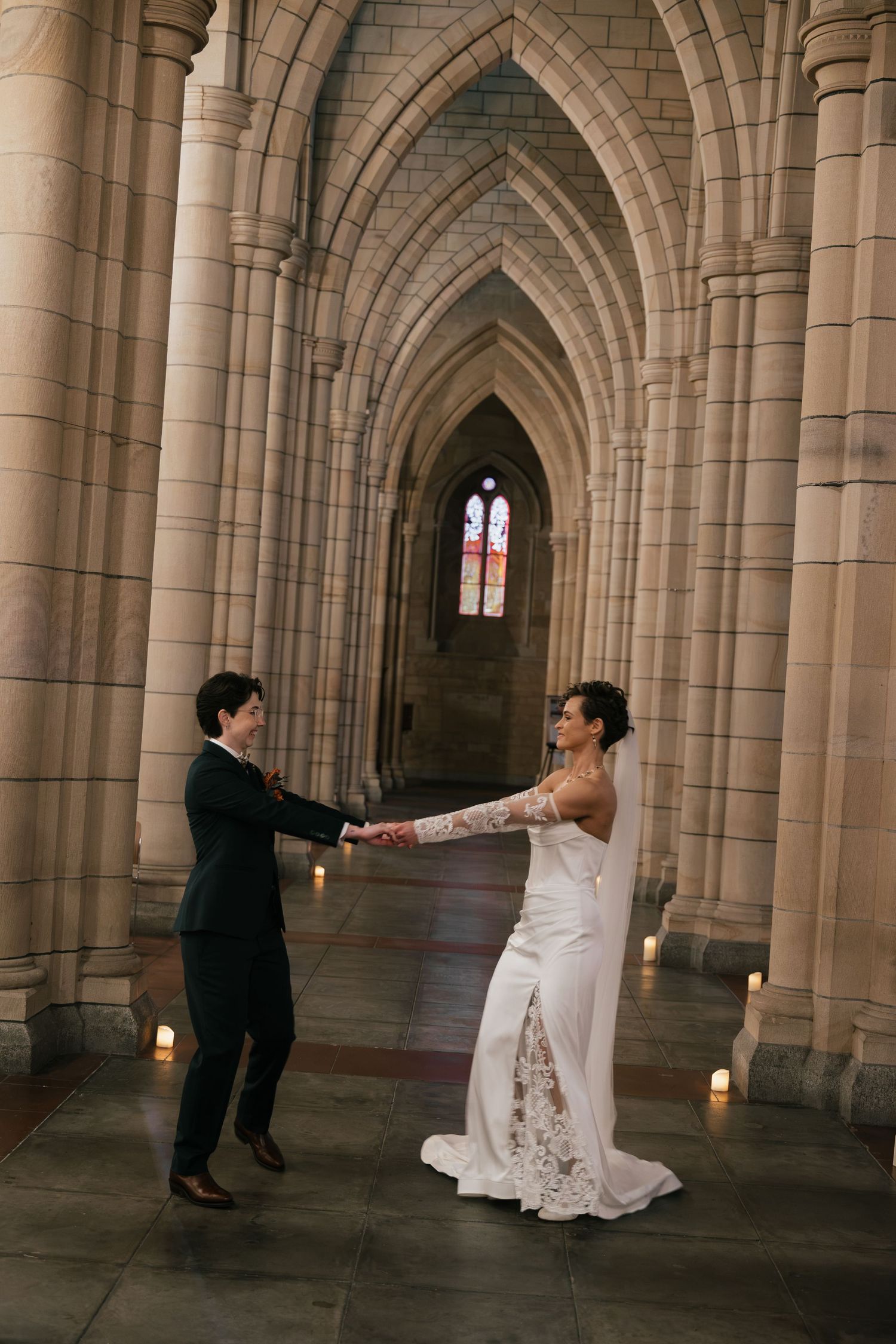 Bride and groom hold hands in gothic cathedral archway, candlelit stone corridor, lace wedding gown, elegant ceremony.