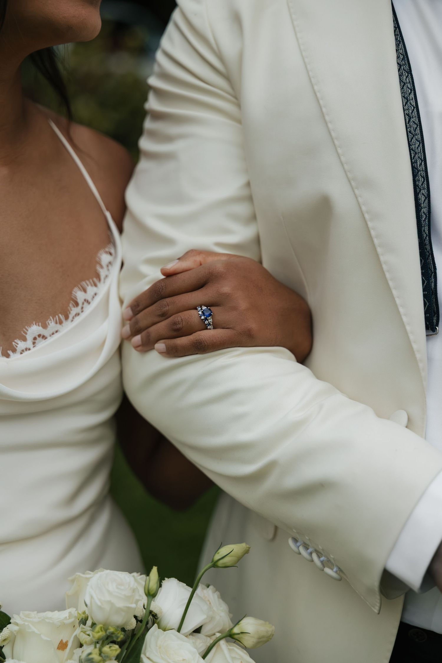 Bride and groom in white attire embrace, showcasing a sapphire ring, with a white rose bouquet.