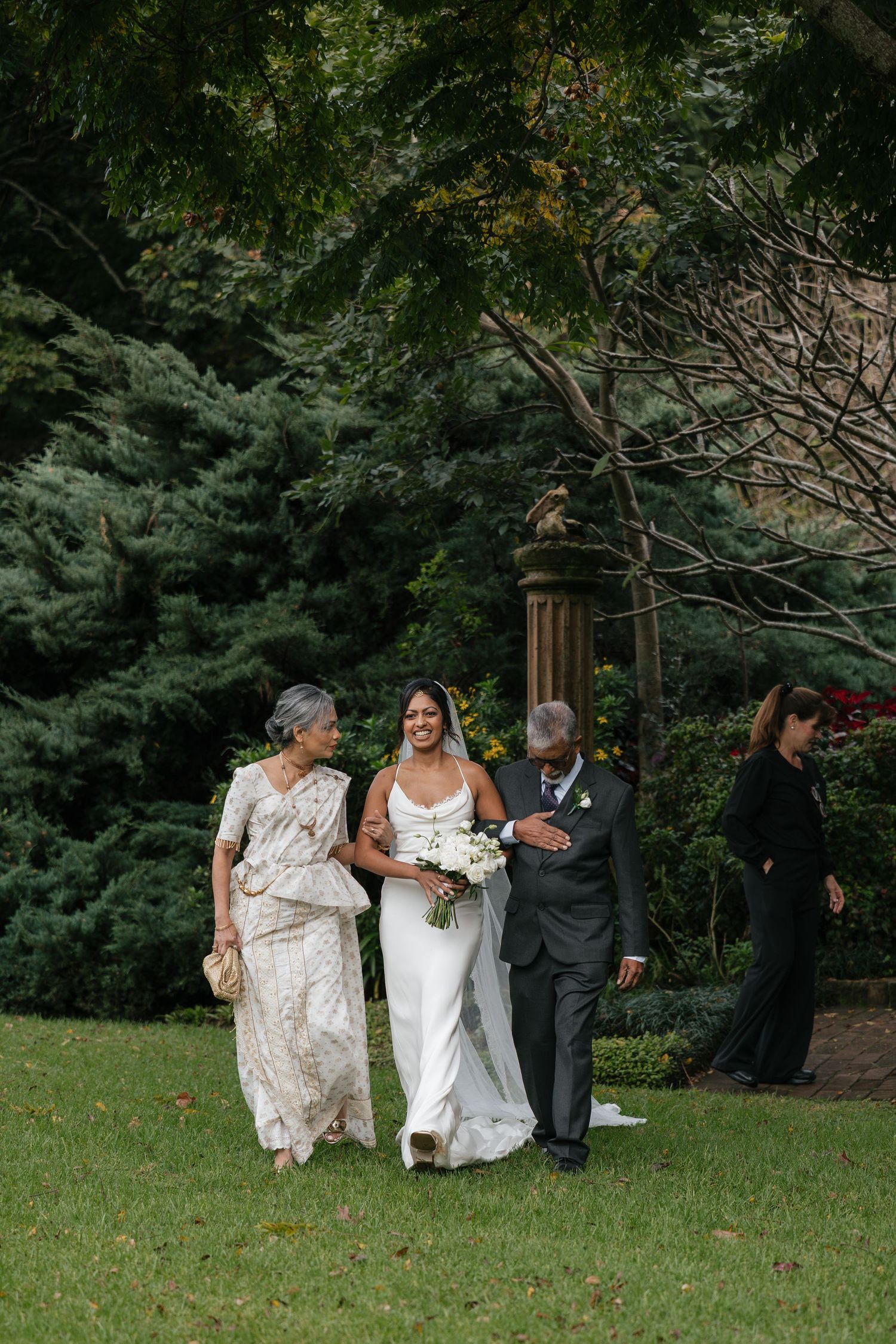 Bride in white gown walks with parents through garden on wedding day, holding white bouquet.