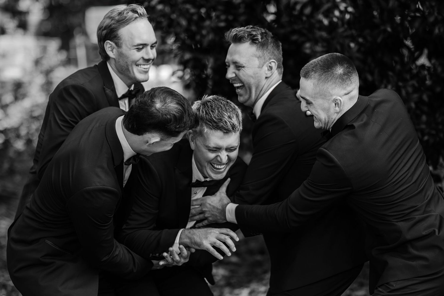 Groomsmen in black suits laughing and playfully hugging the groom outdoors in a candid black and white wedding photo.