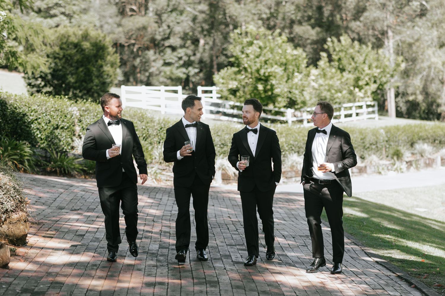 Four groomsmen in black tuxedos and bow ties walking on a brick path outdoors at a wedding celebration.
