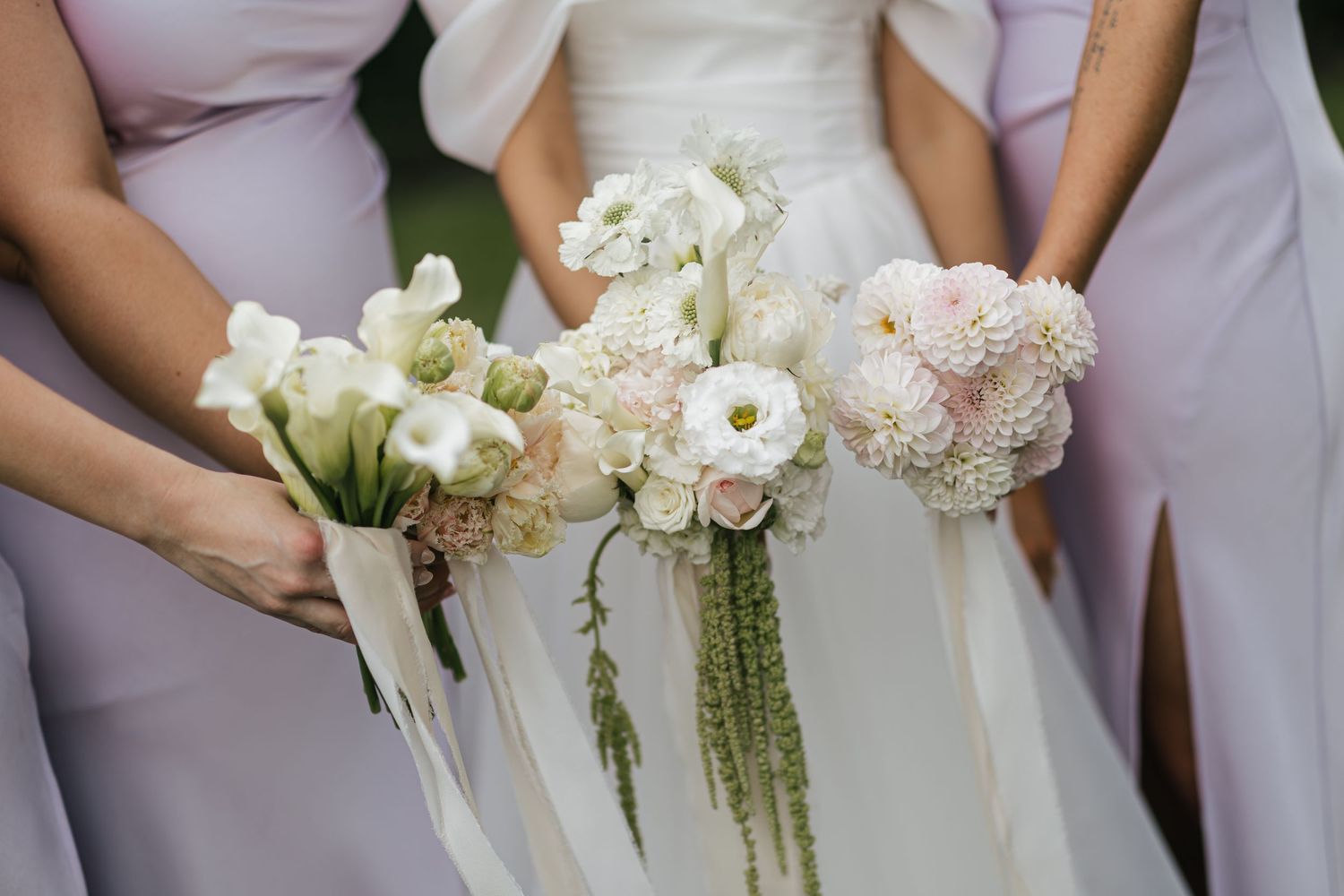 Bride and bridesmaids in lavender dresses holding elegant white and blush pink wedding bouquets with flowing ribbons.