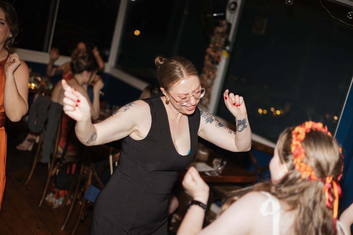Woman with tattoos and glasses dancing joyfully at an indoor party event in a black dress.