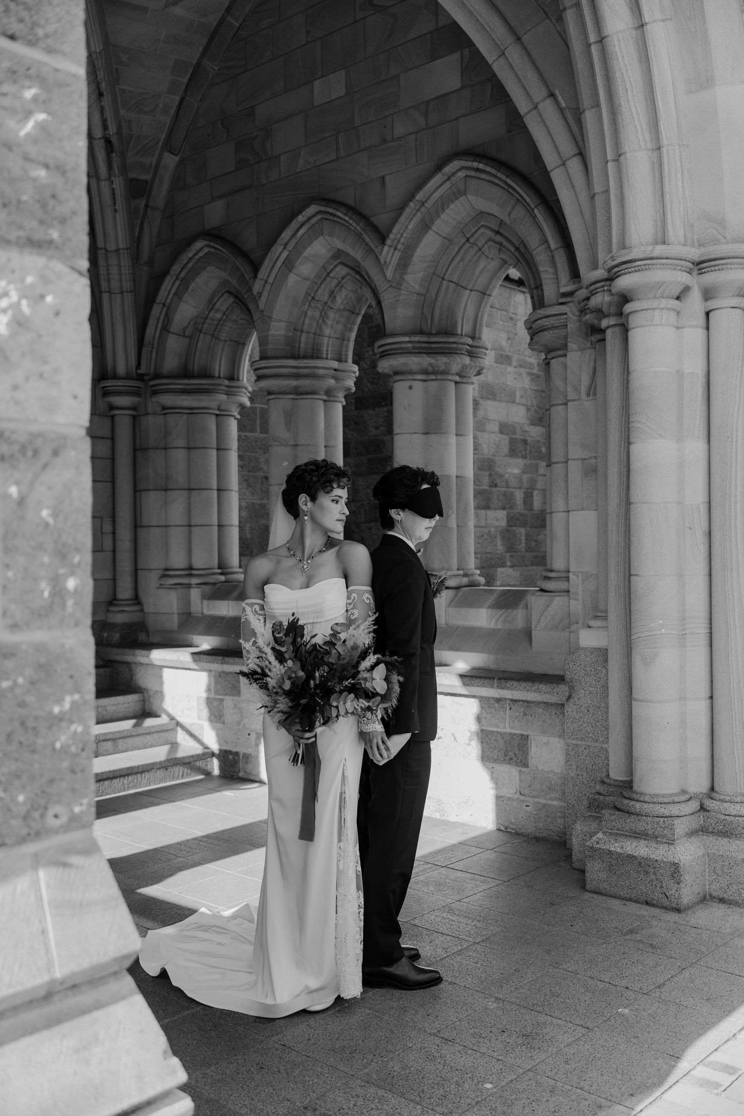 Black and white photo of a bride and groom standing under stone arches at a historic venue.