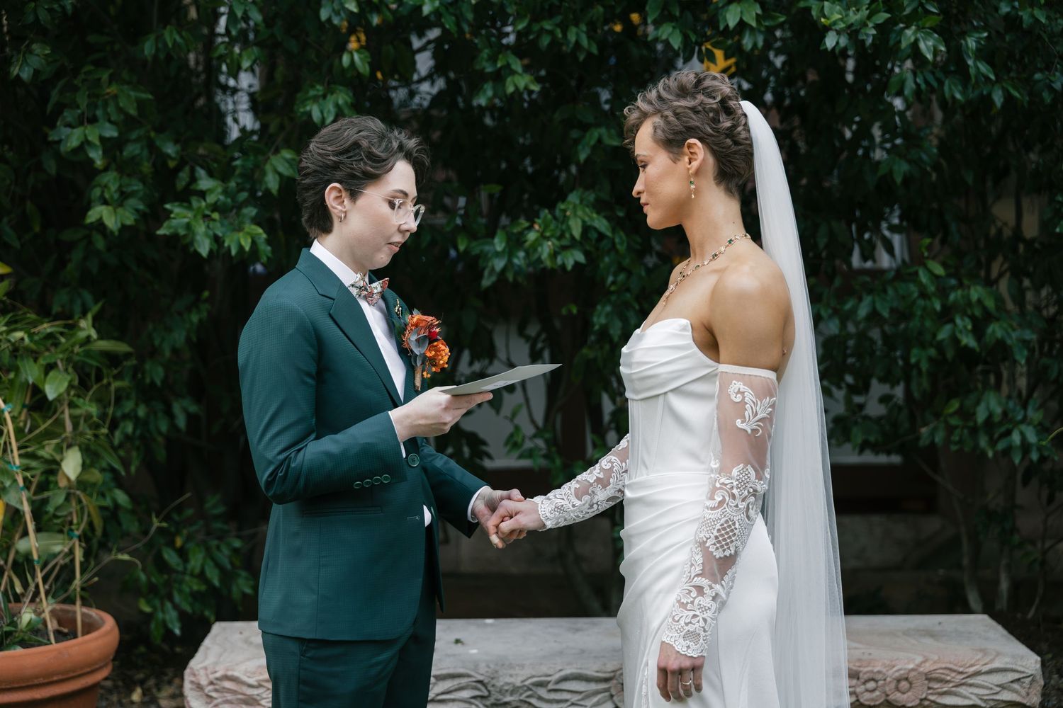 Two brides exchange vows outdoors, one in a teal suit and one in a white lace gown with veil, surrounded by greenery.
