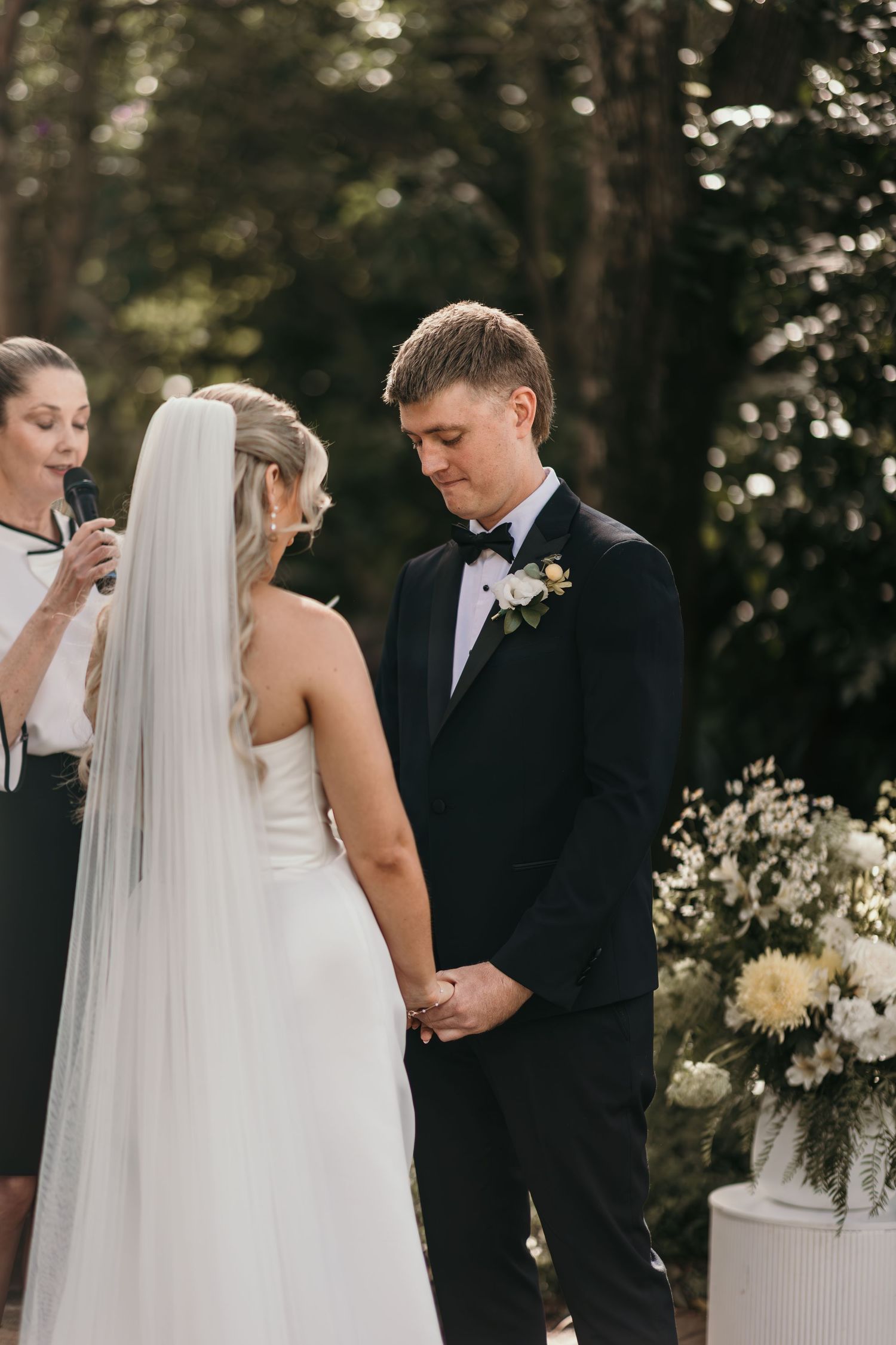 Bride and groom holding hands at outdoor wedding ceremony, officiant in background, white floral arrangements nearby.