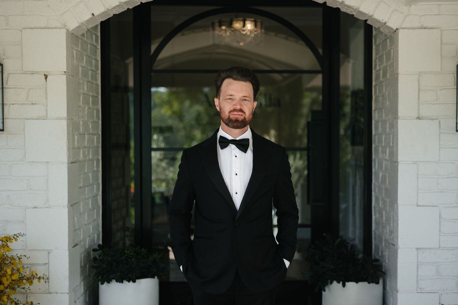 A groom in a black tuxedo and bow tie stands confidently at an elegant arched doorway of a white stone venue.