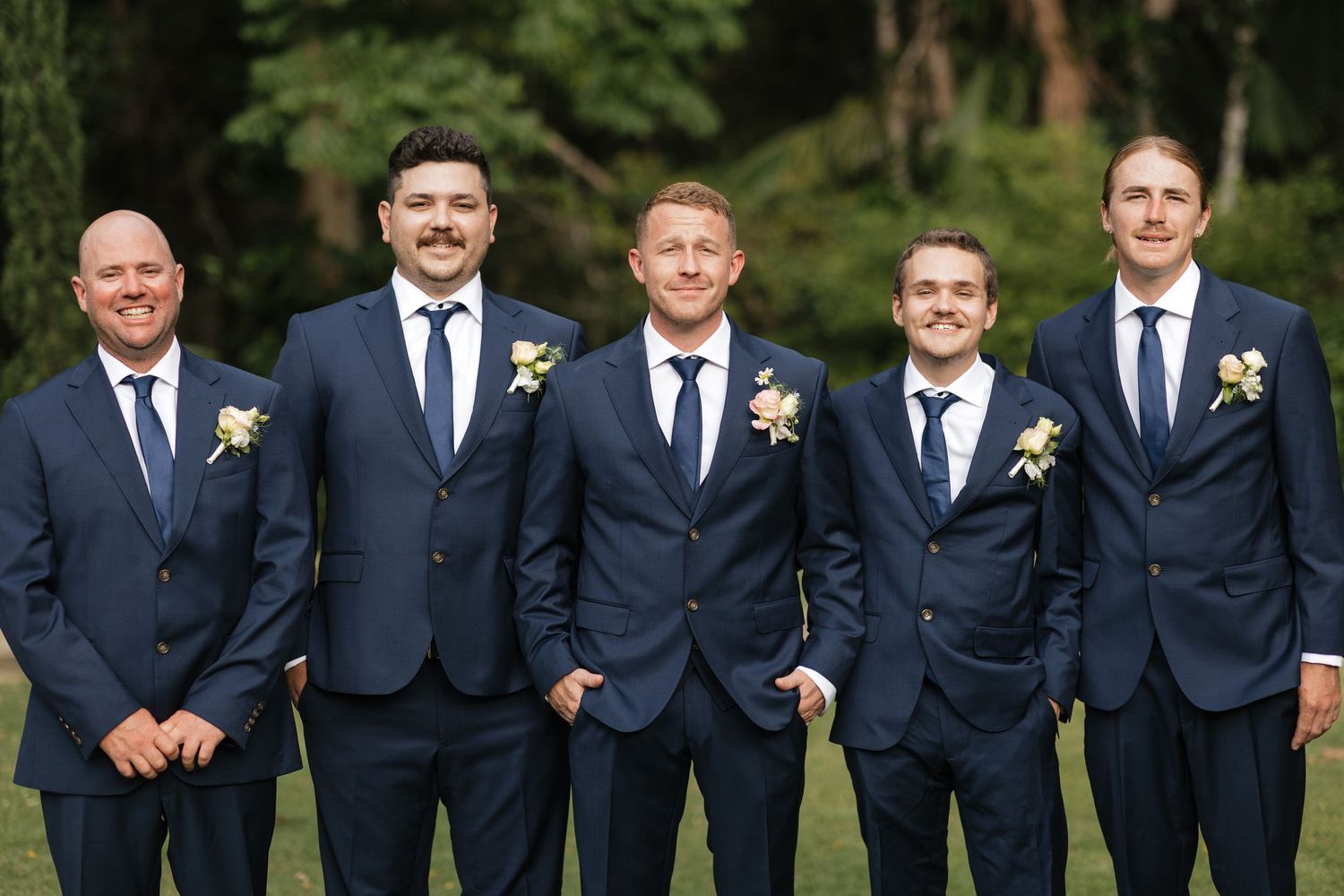 Five groomsmen in matching navy blue suits with boutonnieres posing outdoors for a wedding photo.