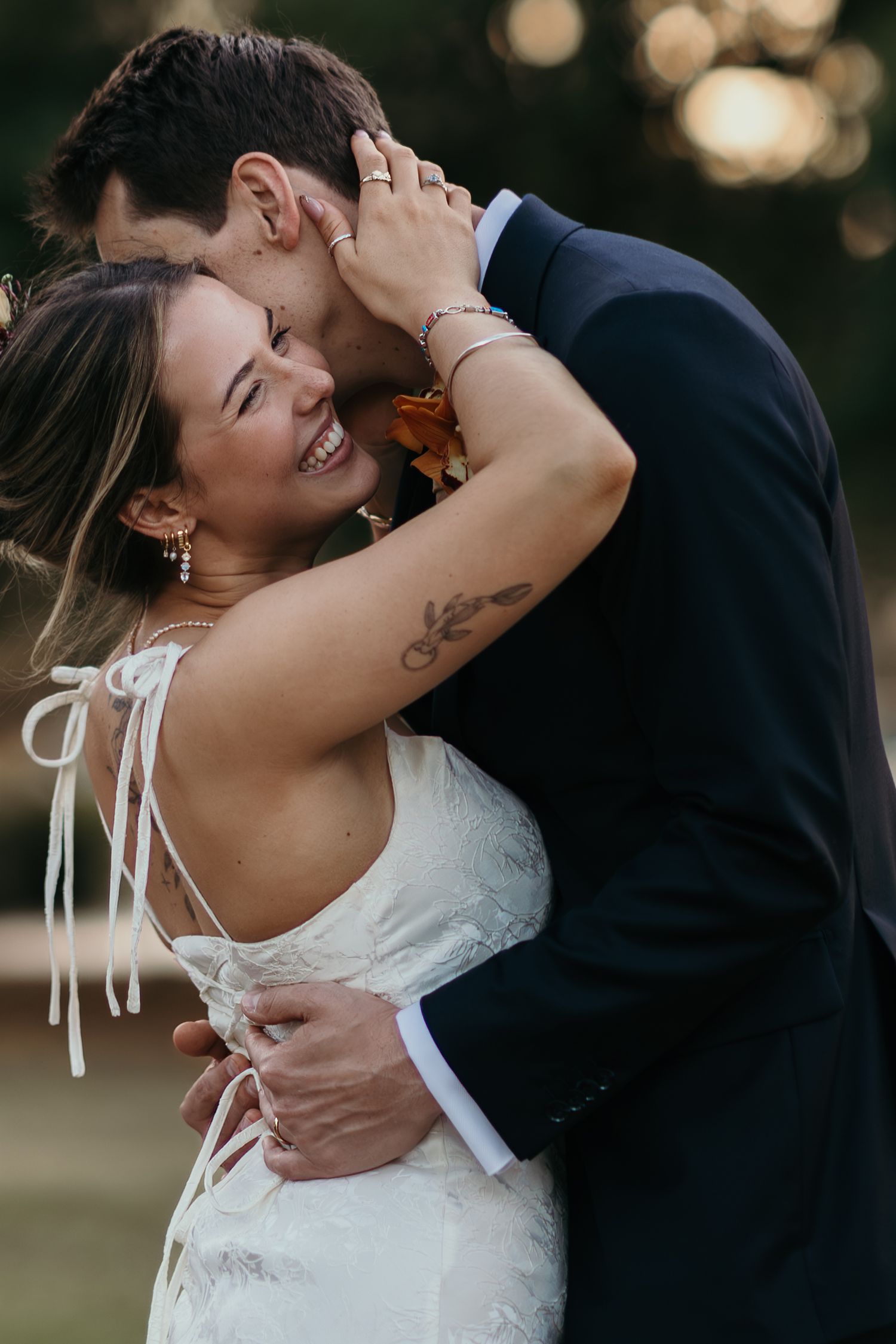 A romantic wedding couple embraces outdoors during golden hour with soft bokeh lighting in the background.
