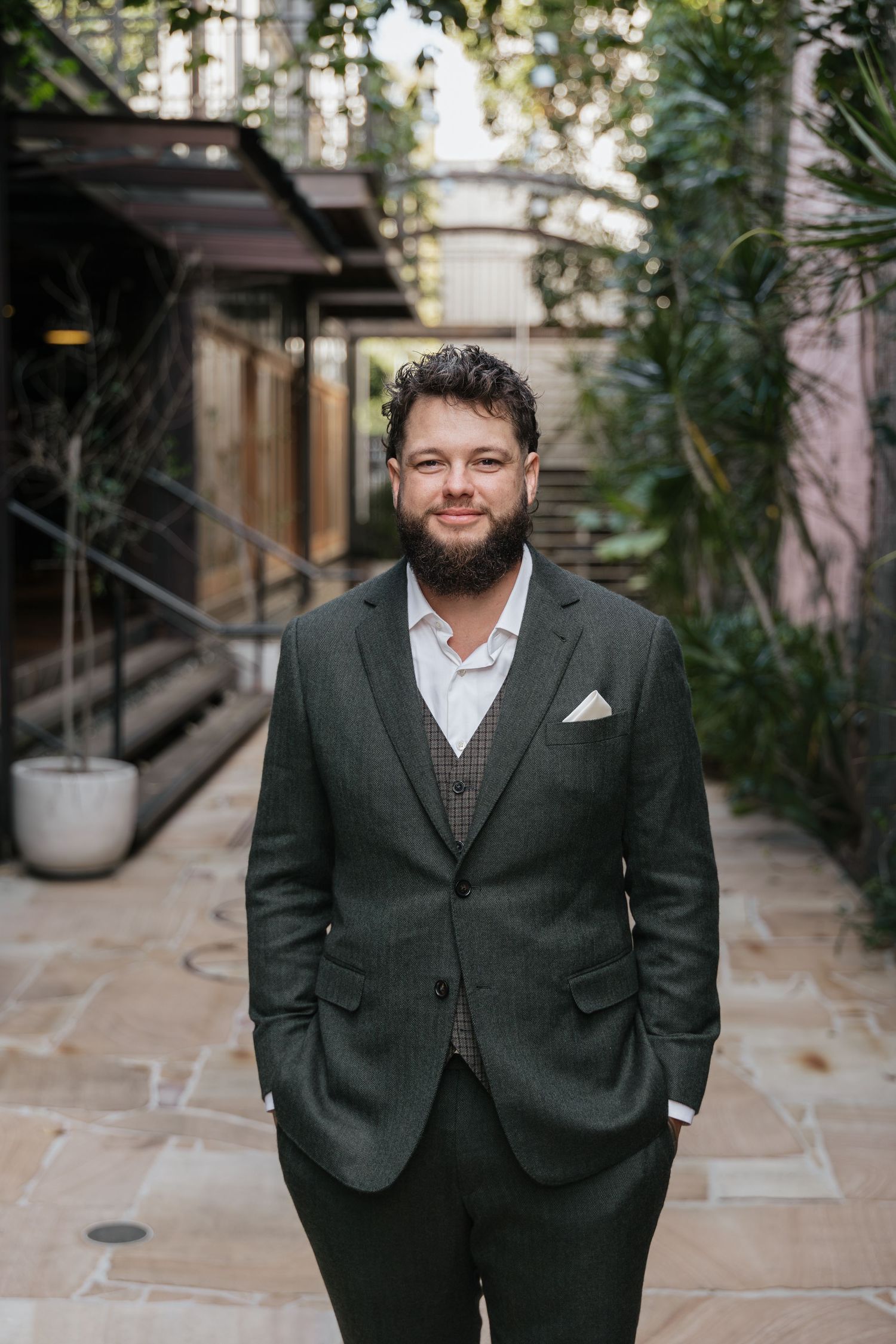 Bearded man in dark gray suit with white pocket square smiling in lush courtyard setting.