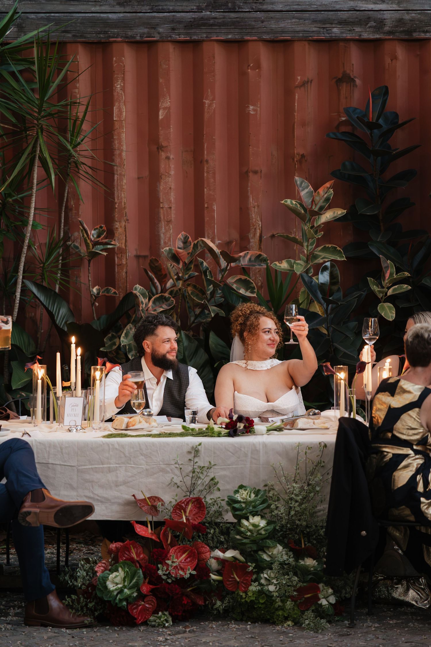 Bride and groom toast at candlelit outdoor wedding reception table surrounded by lush greenery and red floral arrangements.