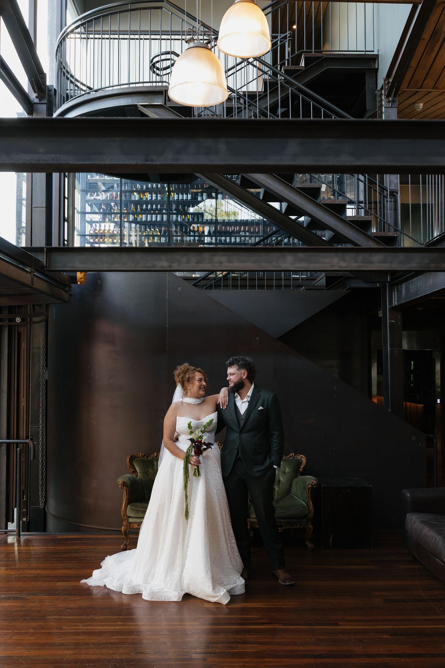 Bride and groom share a romantic moment in an industrial wedding venue with spiral staircase and warm pendant lighting.
