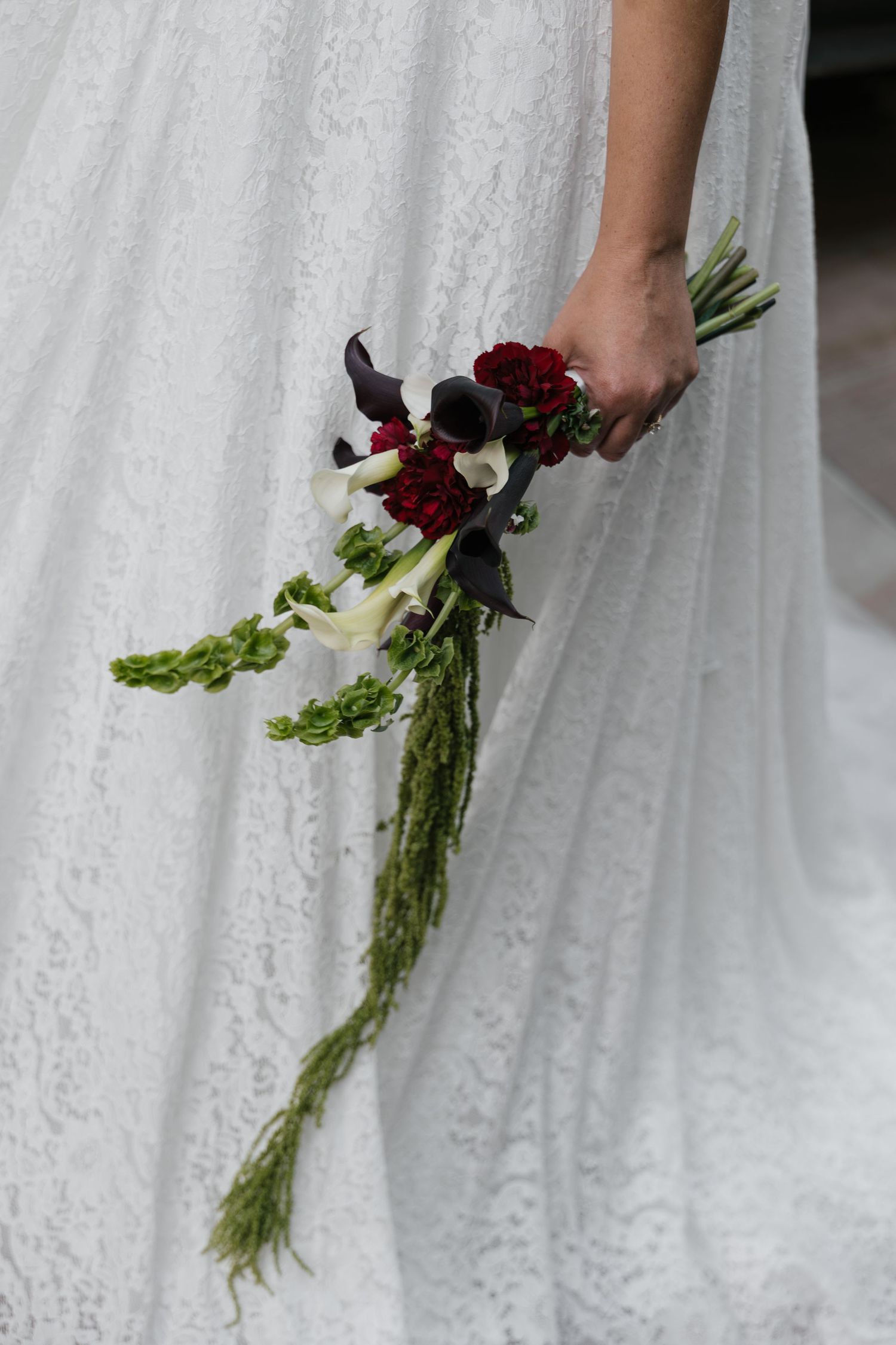 Bride in white lace gown holding cascading bouquet with dark calla lilies, red roses, and trailing greenery.
