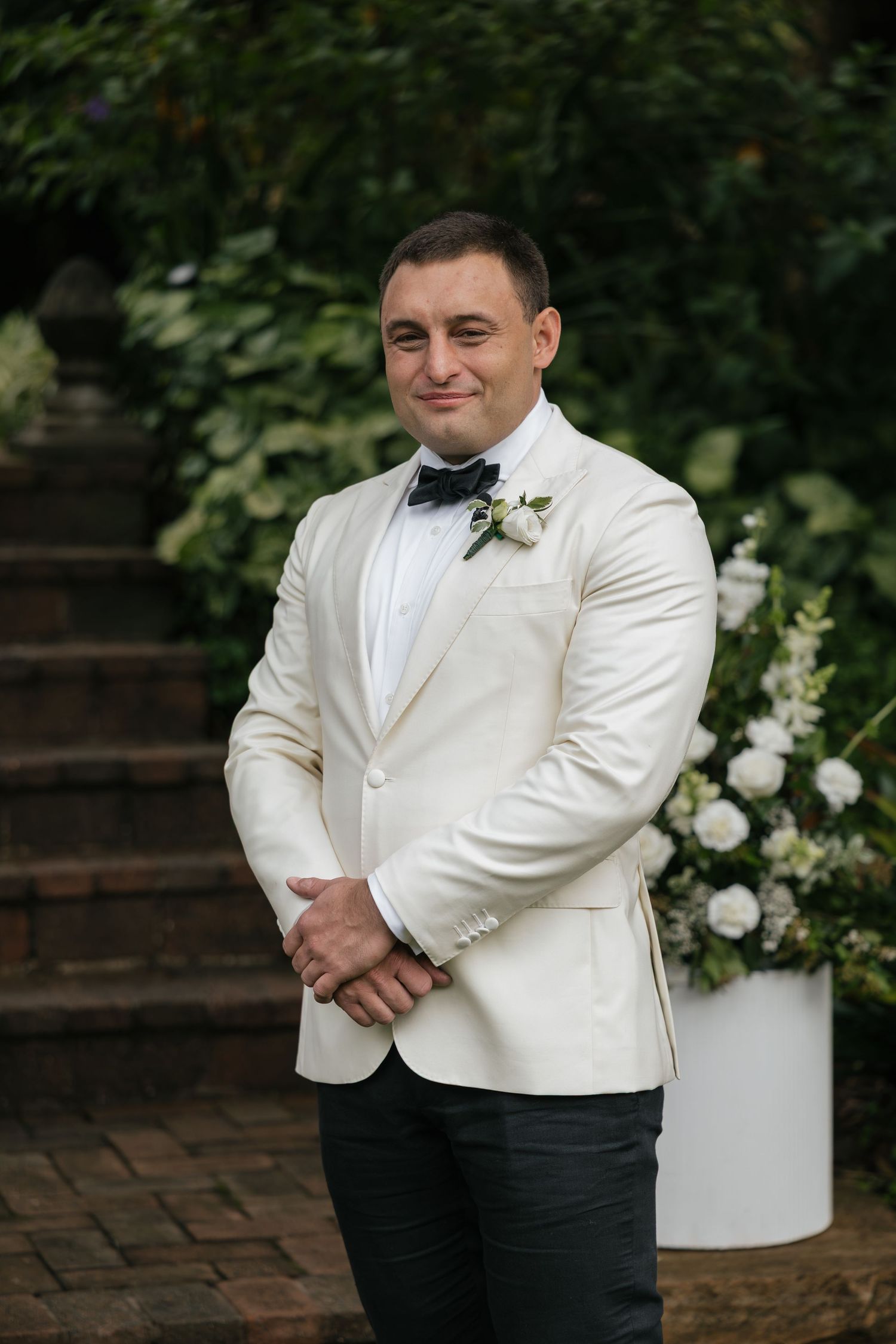 Groom in white tuxedo jacket with black bow tie and boutonniere smiling at outdoor wedding ceremony.