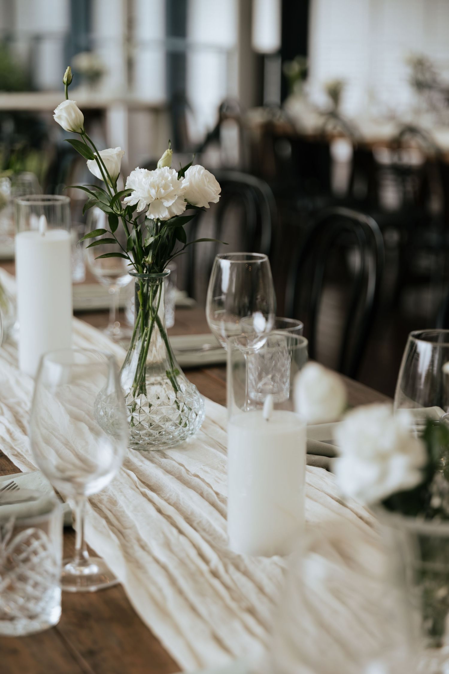 Elegant wedding table setting with white pillar candles, crystal glassware, and white peonies in glass vases on a rustic wood table.