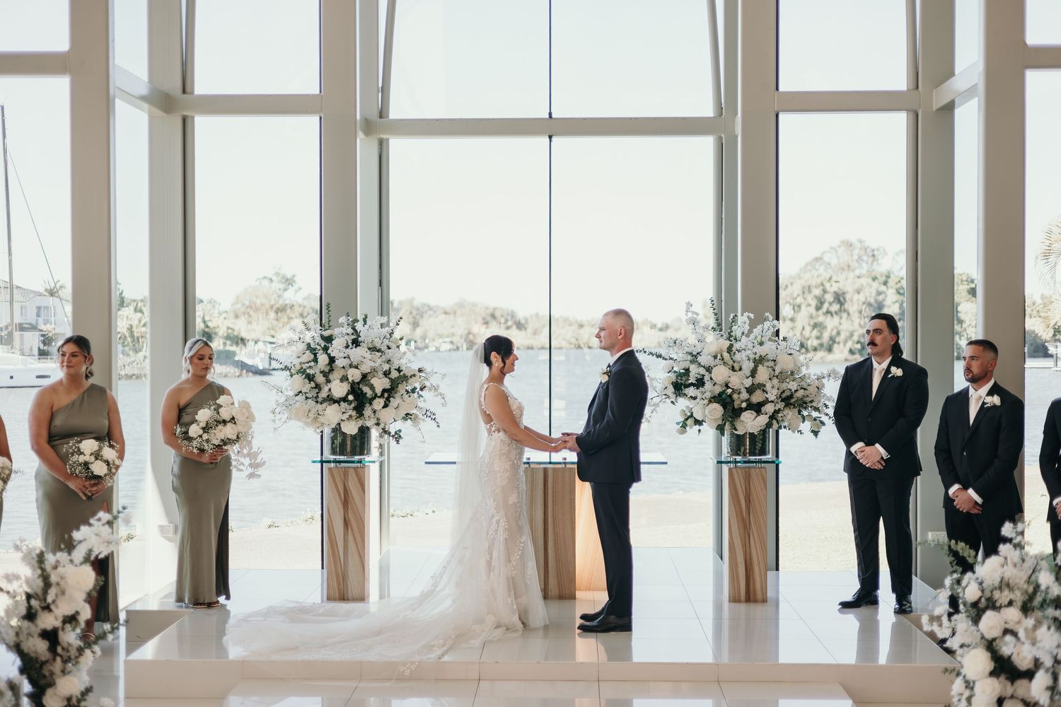 A wedding ceremony takes place in a modern chapel with floor-to-ceiling windows and elegant white floral arrangements.