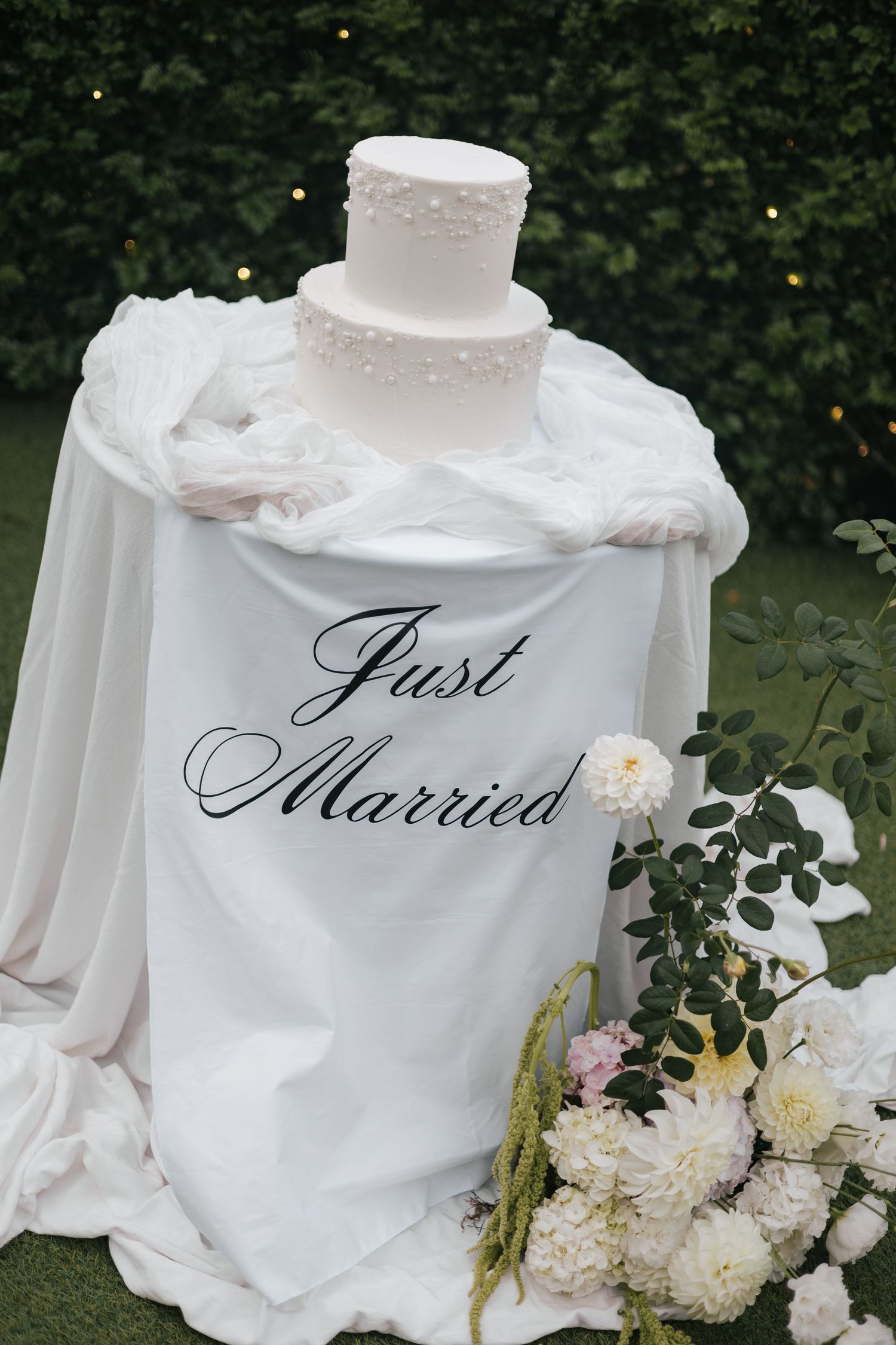 Elegant white wedding cake on a draped table with Just Married sign, surrounded by white flowers outdoors.
