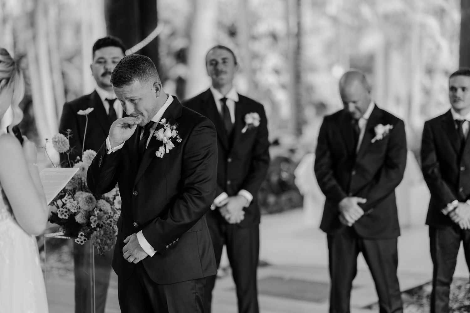 Emotional groom wiping tears as bride approaches at outdoor wedding ceremony with groomsmen standing behind.