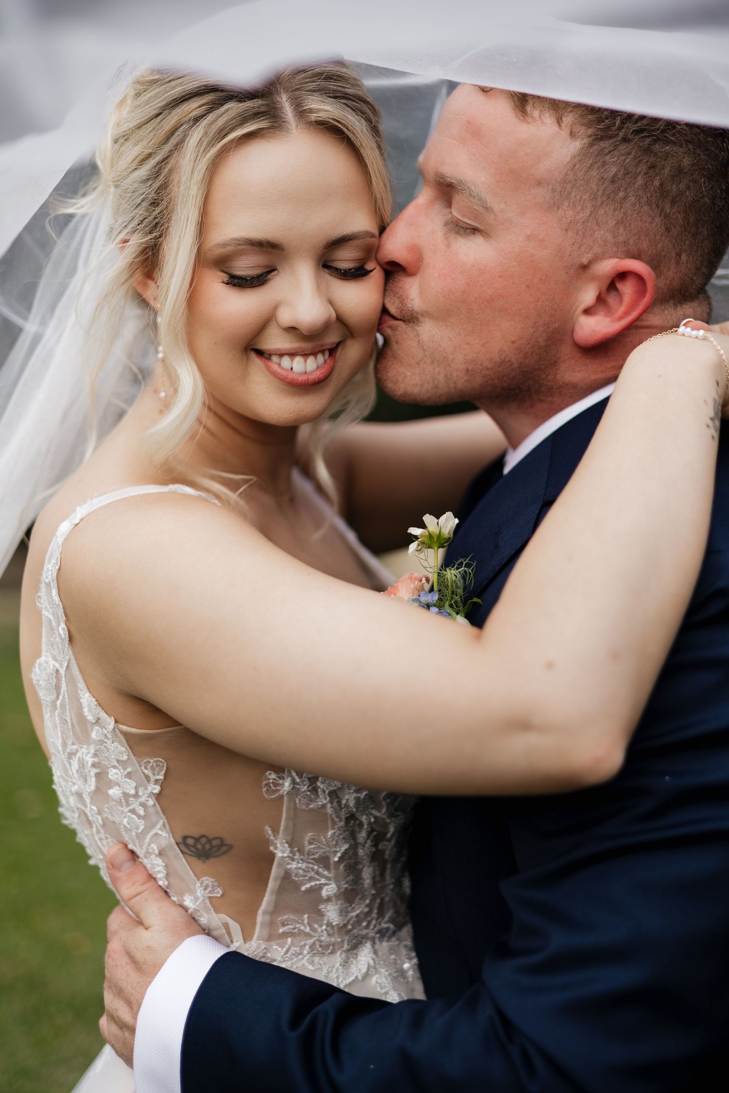 Groom in navy suit kissing bride on cheek as she smiles, both embracing under wedding veil outdoors.